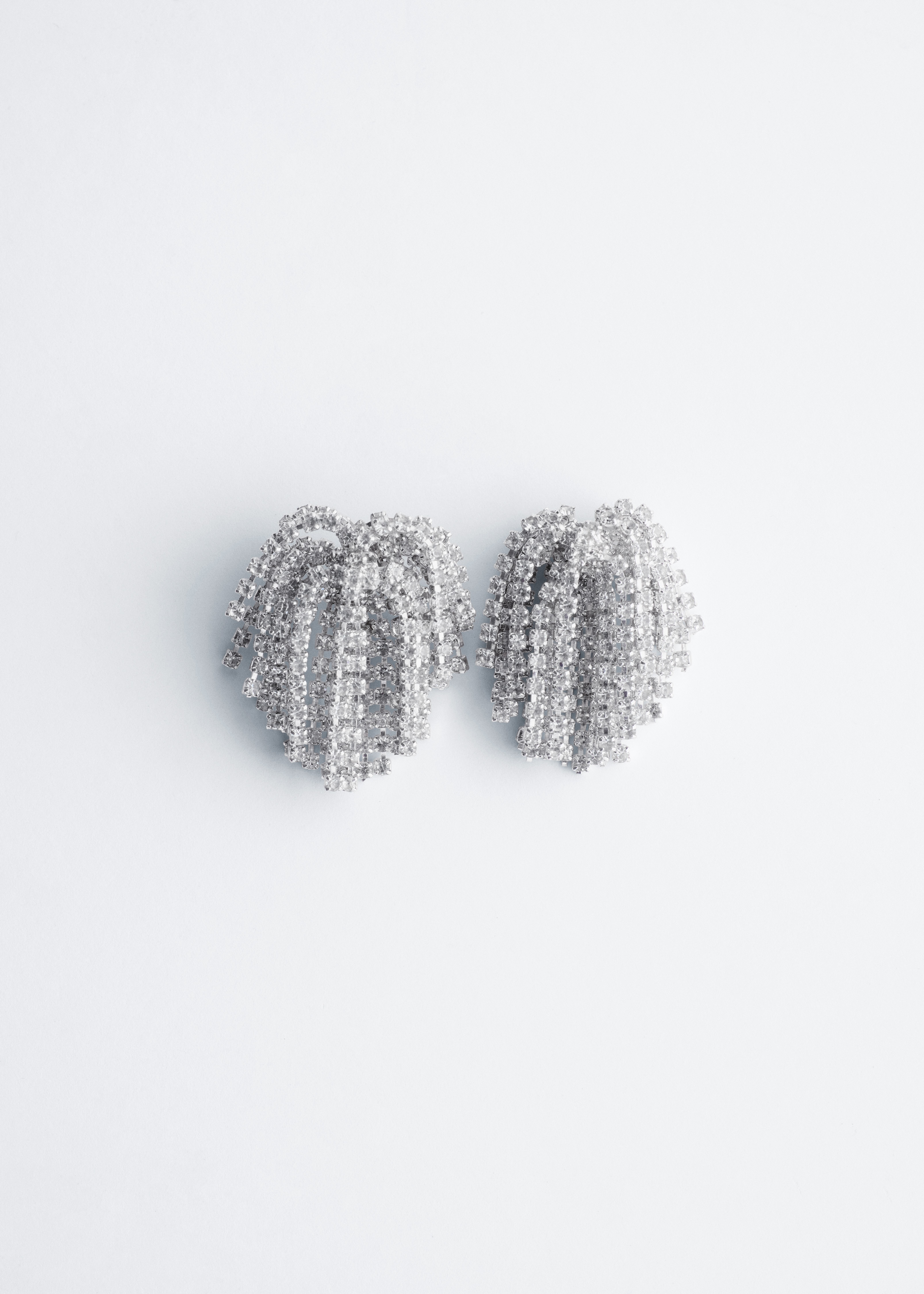 Silver-toned fringe earrings embellished with rhinestones are being worn by the model, paired with a brown and beige patterned satin dress. The dress features a high round neckline and subtle gathers at the shoulders.
