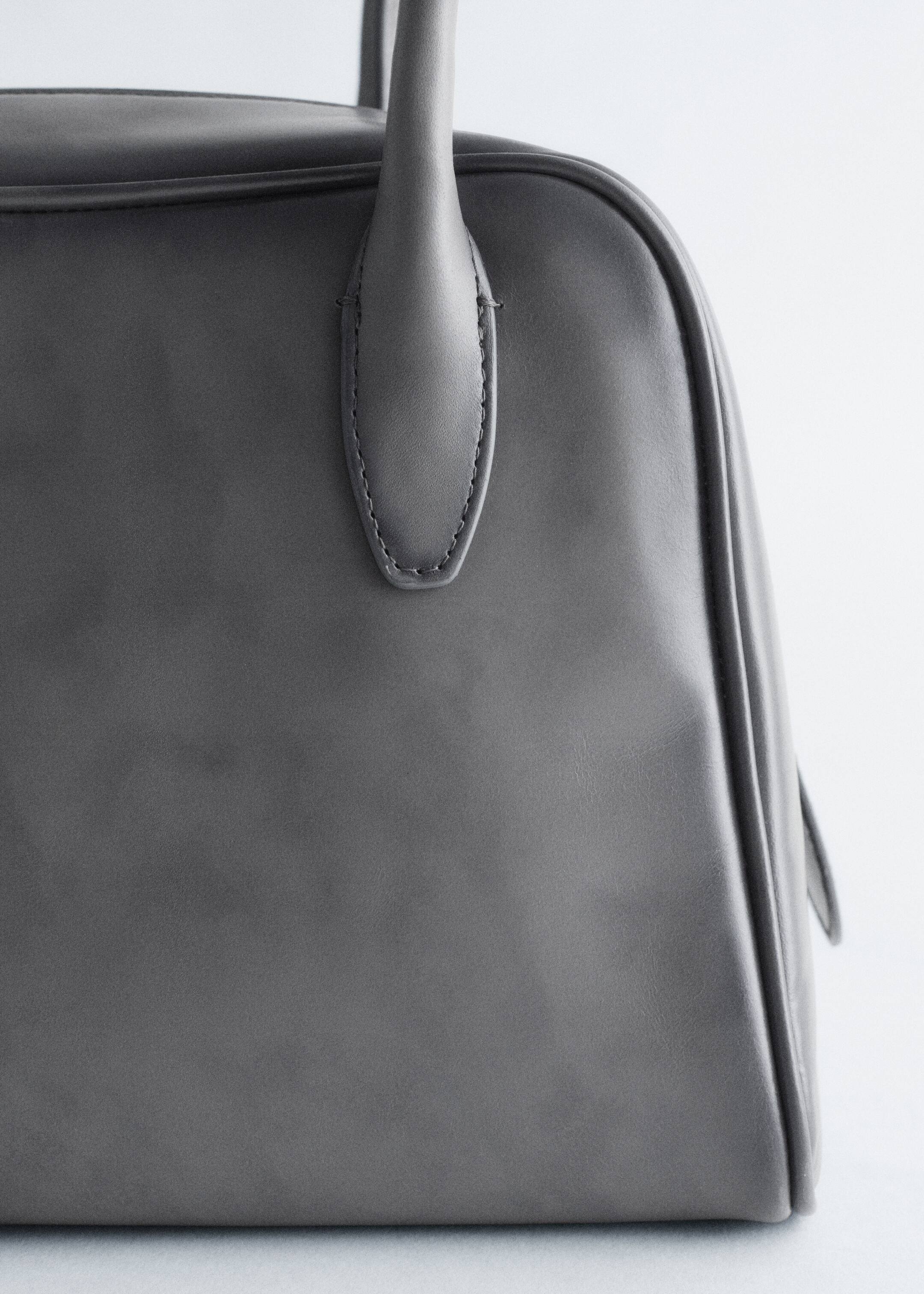 Grösseres Bild ansehen: Eine strukturierte, dunkelgraue Handtasche wird gezeigt, die ein glattes, mattes Finish aufweist. Ein oberer Griff erhebt sich, befestigt am Körper der Tasche mit einem genähten, tropfenförmigen Tab. Der obere Rand der Tasche und ein Teil ihres seitlichen Faltenbalgs sind sichtbar.