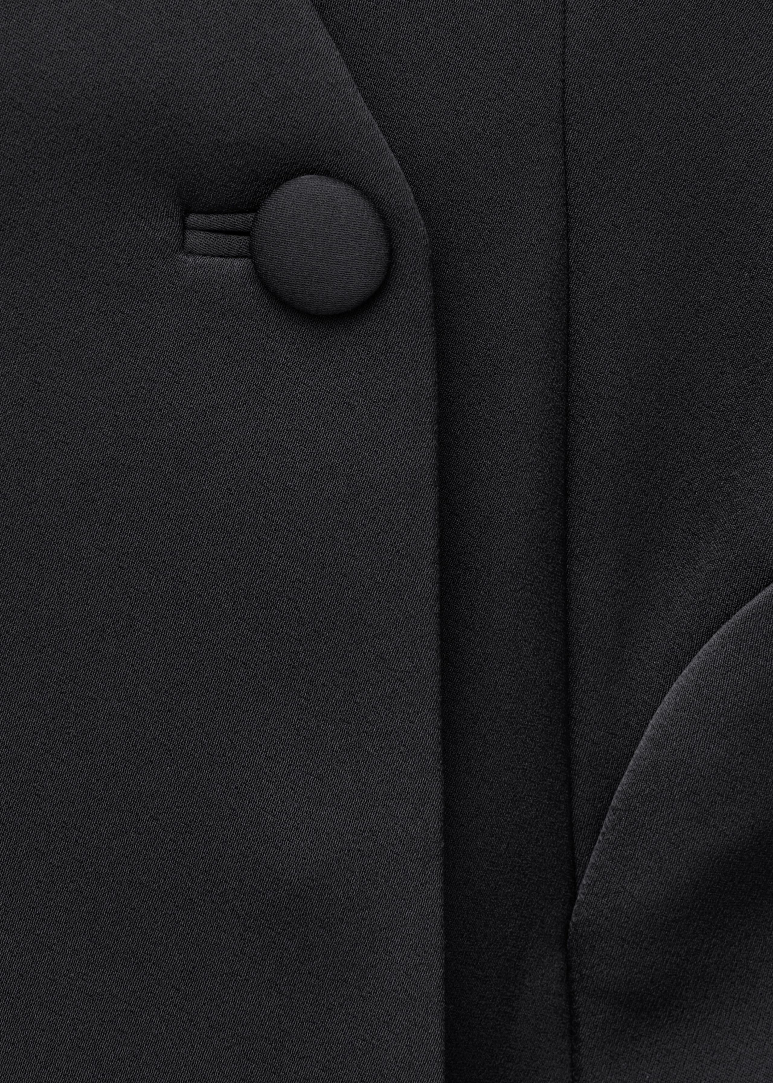 A close-up view of the black blazer dress shows a single, round, fabric-covered button fastened through a buttonhole. The image highlights the fine woven texture of the black fabric and clean vertical seam details.