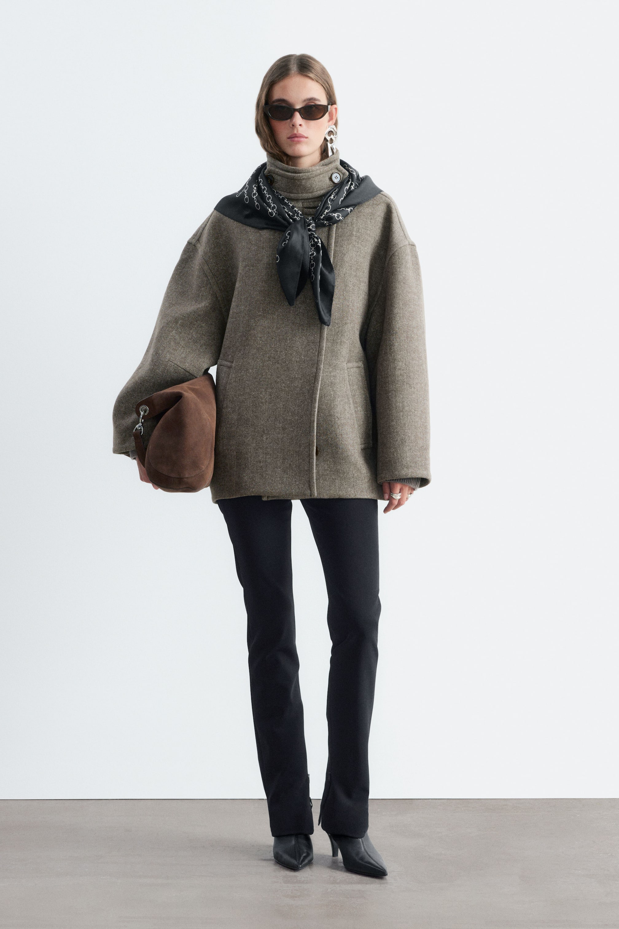 Agrandir l'image: Un manteau taupe-gris texturé à motif chevron, à la silhouette oversize, aux larges manches raglan et doté d’un col montant à patte est porté par le mannequin. Une écharpe noire à motifs est nouée autour du col du manteau, et un sac en daim brun est porté sous le bras du mannequin.