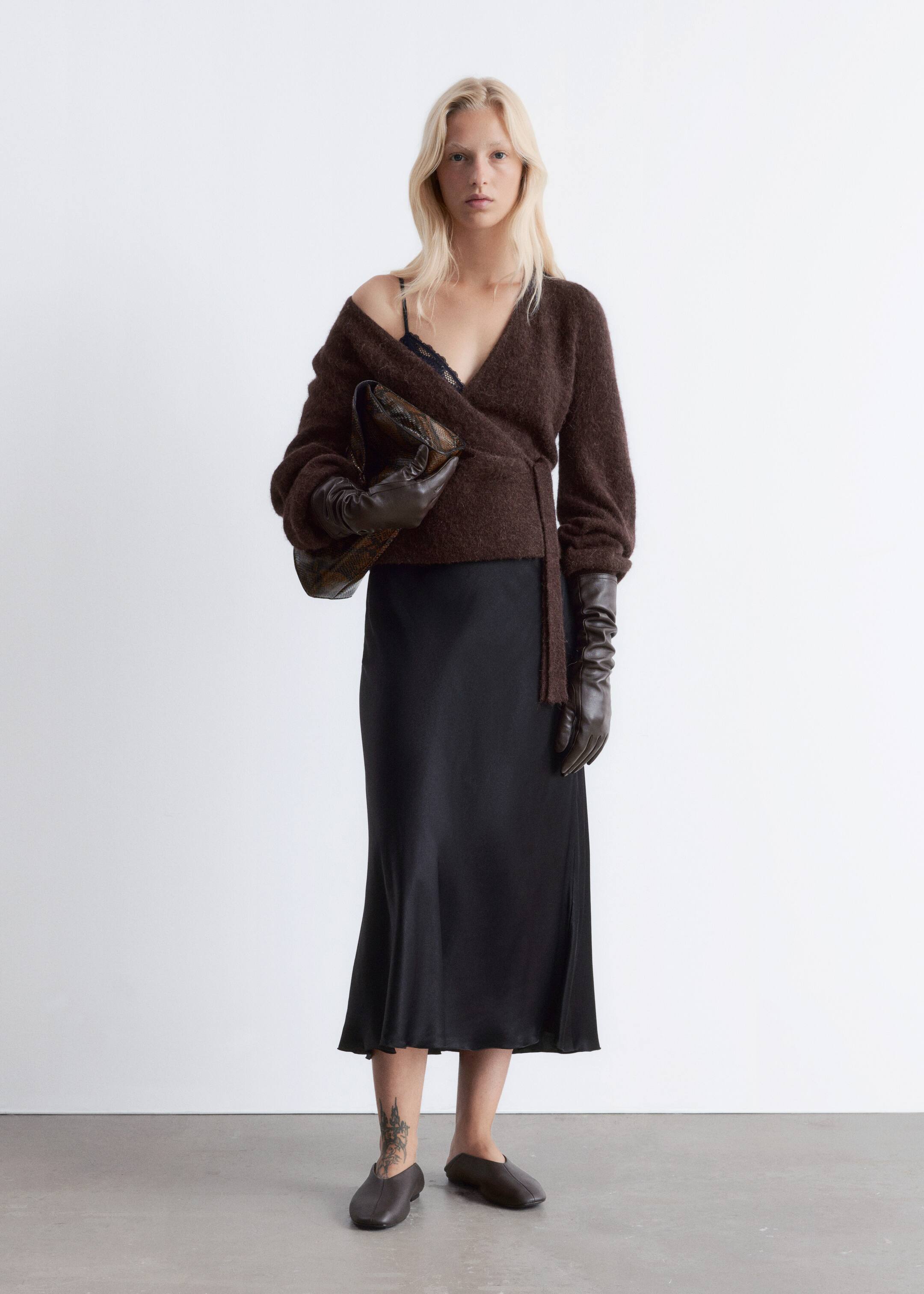 Ingrandisci l'immagine: Un cardigan avvolgente marrone scuro e peloso, con maniche a sbuffo, polsini a coste e chiusura con cintura, è indossato dalla modella. Una spalla del cardigan è portata giù, rivelando un reggiseno a triangolo in pizzo nero, e lunghi guanti marroni sono indossati, completando il cardigan.