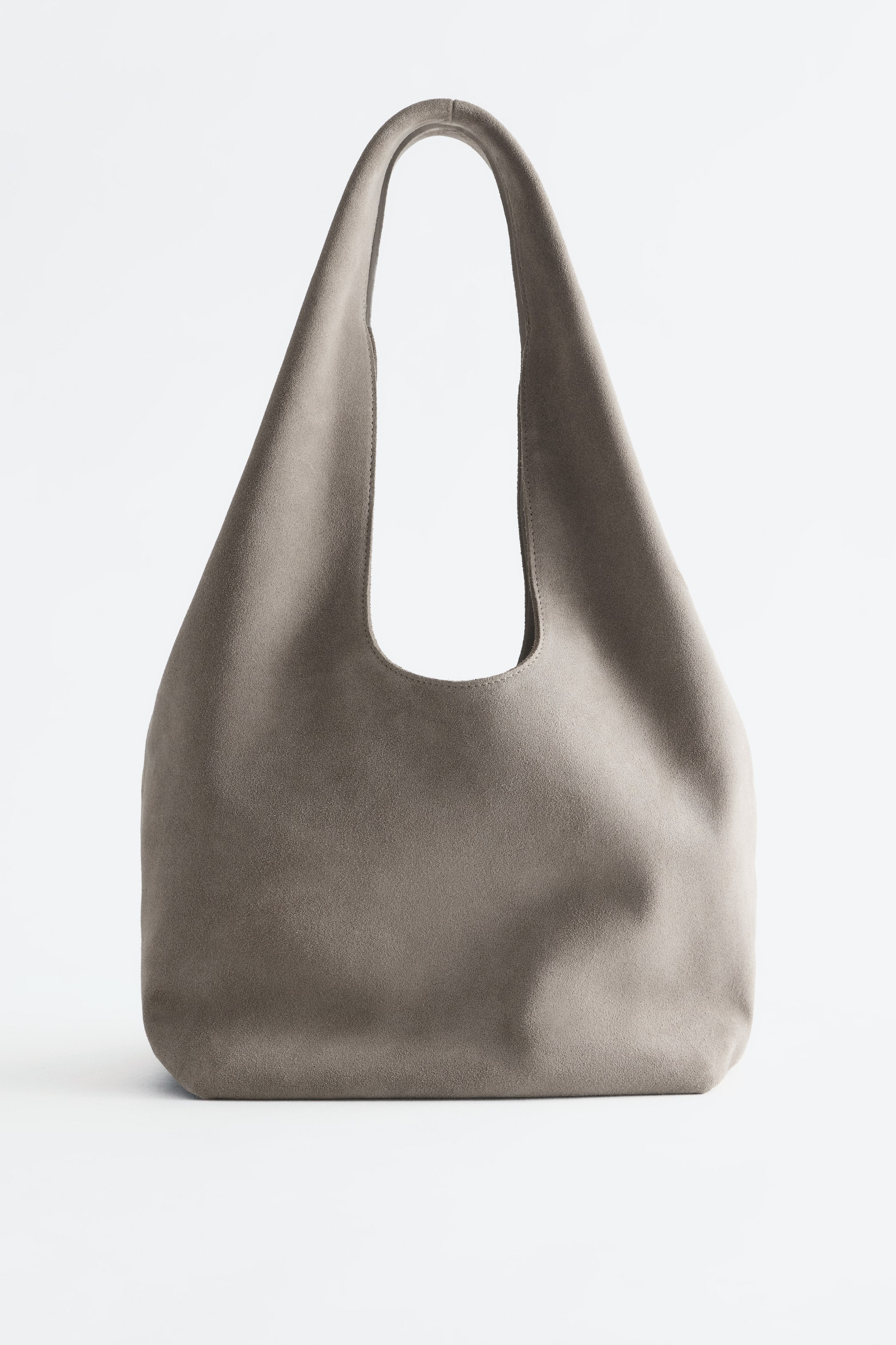 Vis større bilde: En todelt totebag i semsket skinn i en taupegrå nyanse står oppreist, vendt forfra. Den viser en slouchy, romslig silhuett med en bred, integrert skulderrem og flat bunn.
