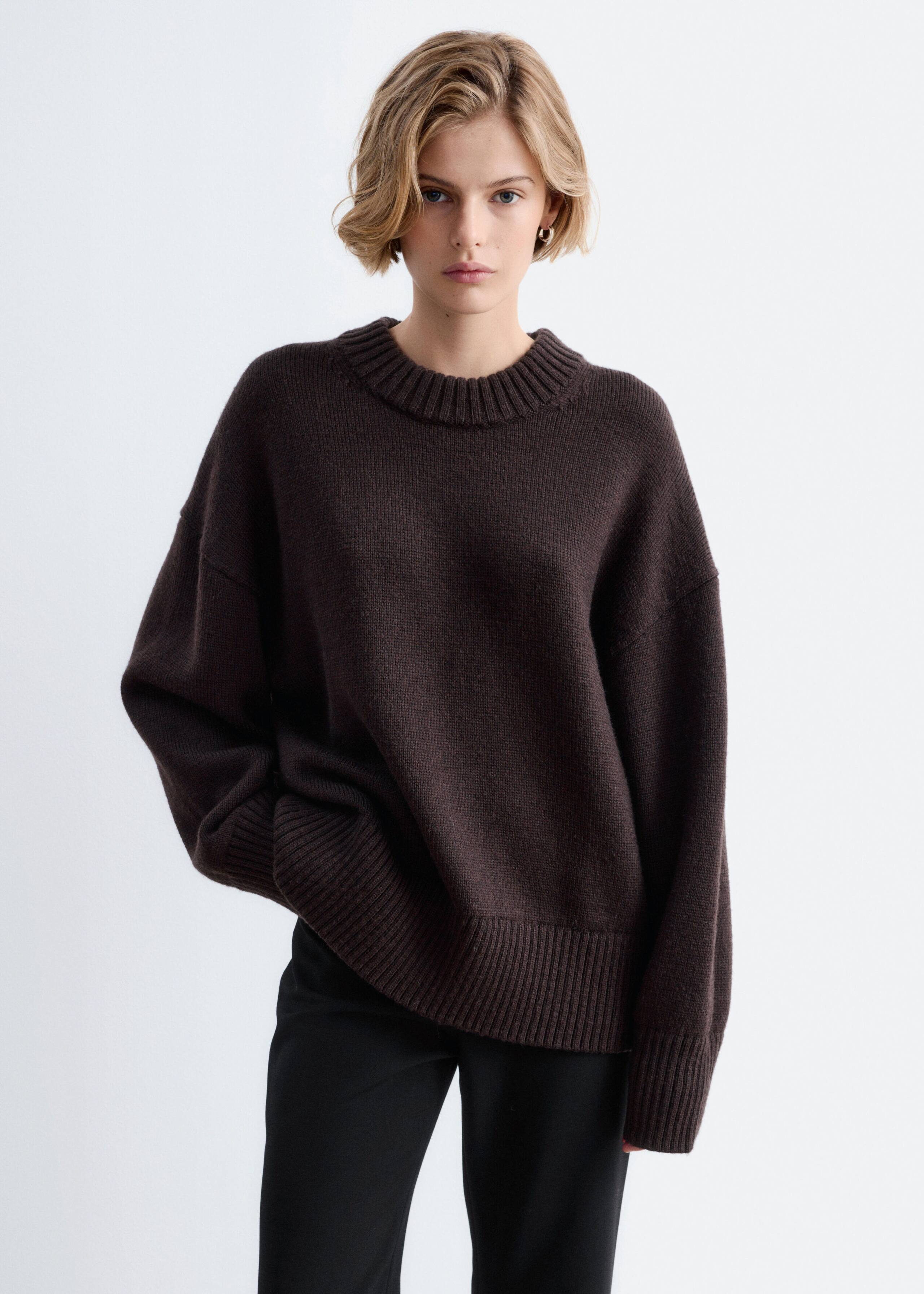 The model is wearing an oversized dark brown wool jumper featuring a ribbed crew neck, dropped shoulders, and wide ribbed cuffs and hem. The jumper is styled with black trousers, with the model resting one hand beneath the hem.