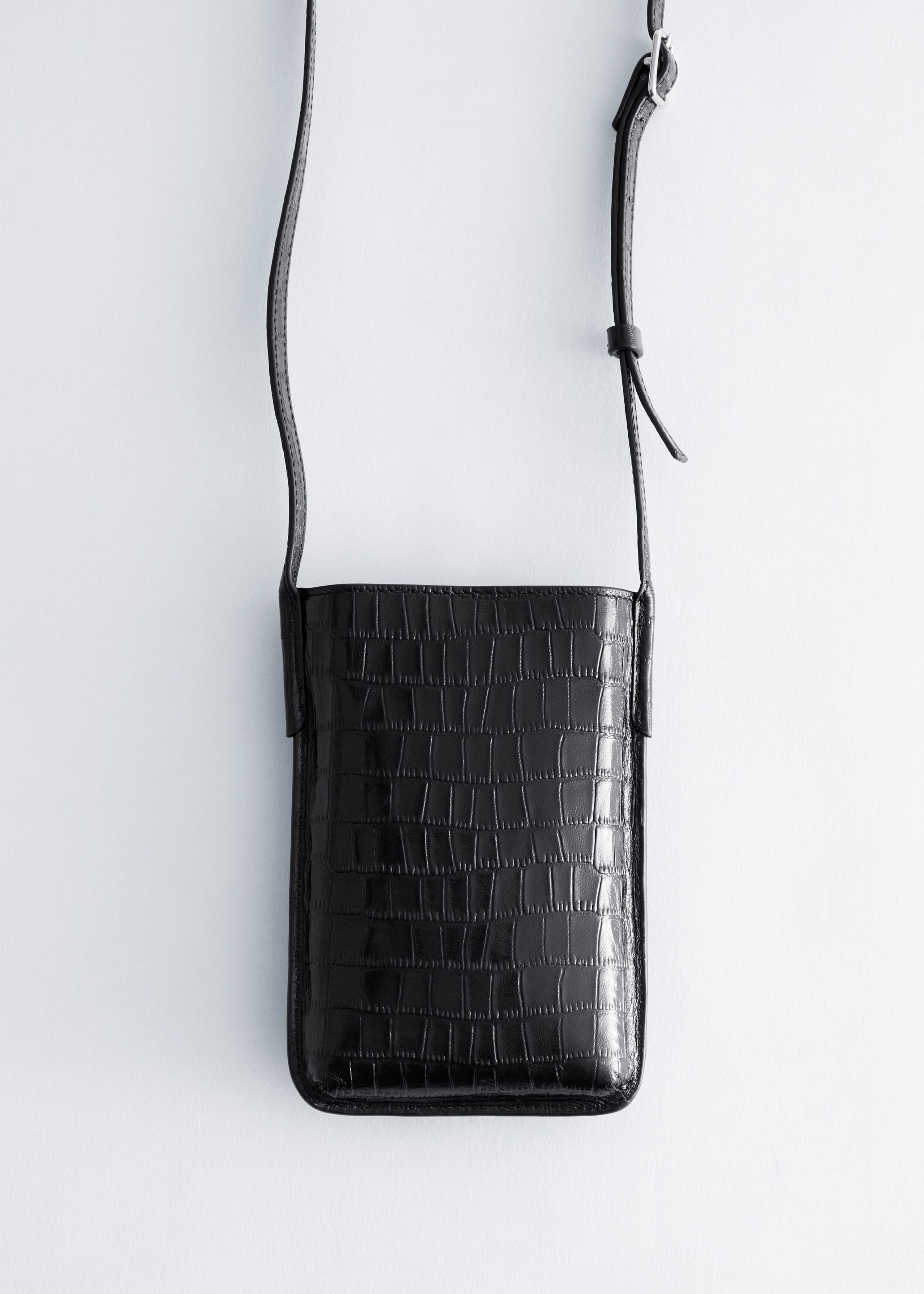 Un sac à téléphone noir, allongé, présentant une texture embossée façon crocodile, est suspendu et montre son design de face. Il adopte une forme rectangulaire aux coins inférieurs arrondis et est porté par une fine bandoulière réglable dotée d’une petite boucle argentée.
