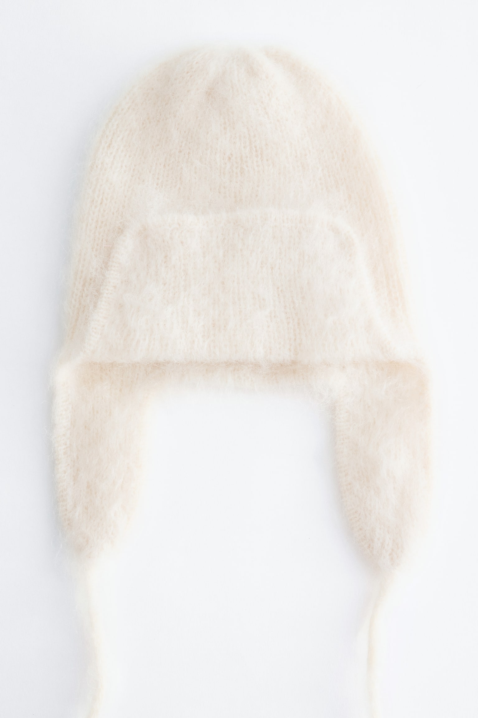 En krämfärgad stickad trapperhatt är vänd framåt och visar upp en fluffig, borstad struktur. Designen har sidolappar för öronen med tunna hängande hakband och en uppvikt frontflik.
