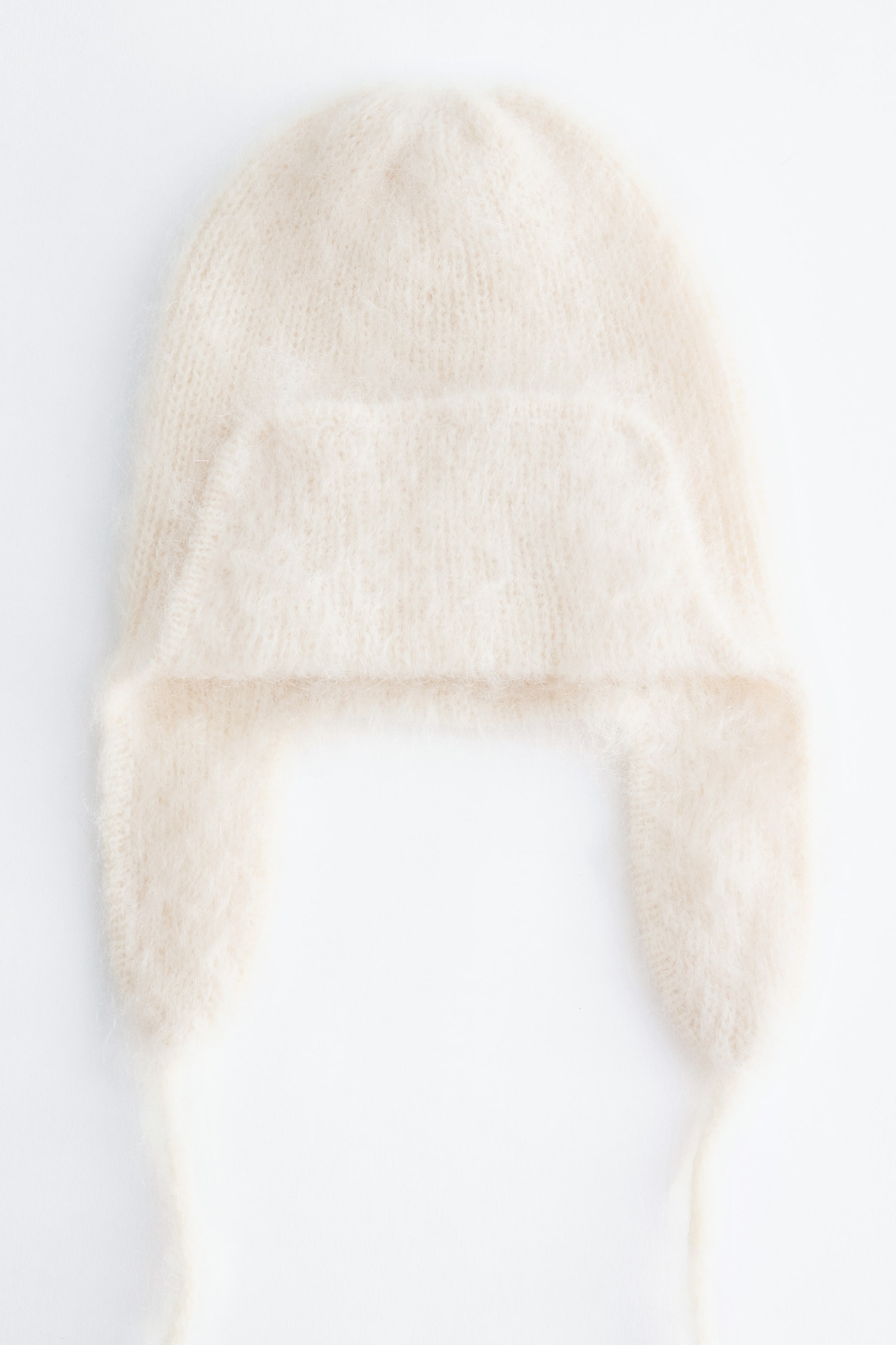 Visualizza immagine più grande: Un cappello trapper in maglia color crema è ripreso frontalmente, mettendo in risalto una texture soffice e spazzolata. Il design presenta paraorecchie laterali con sottili lacci sottogola pendenti e una falda frontale ripiegata verso l’alto.