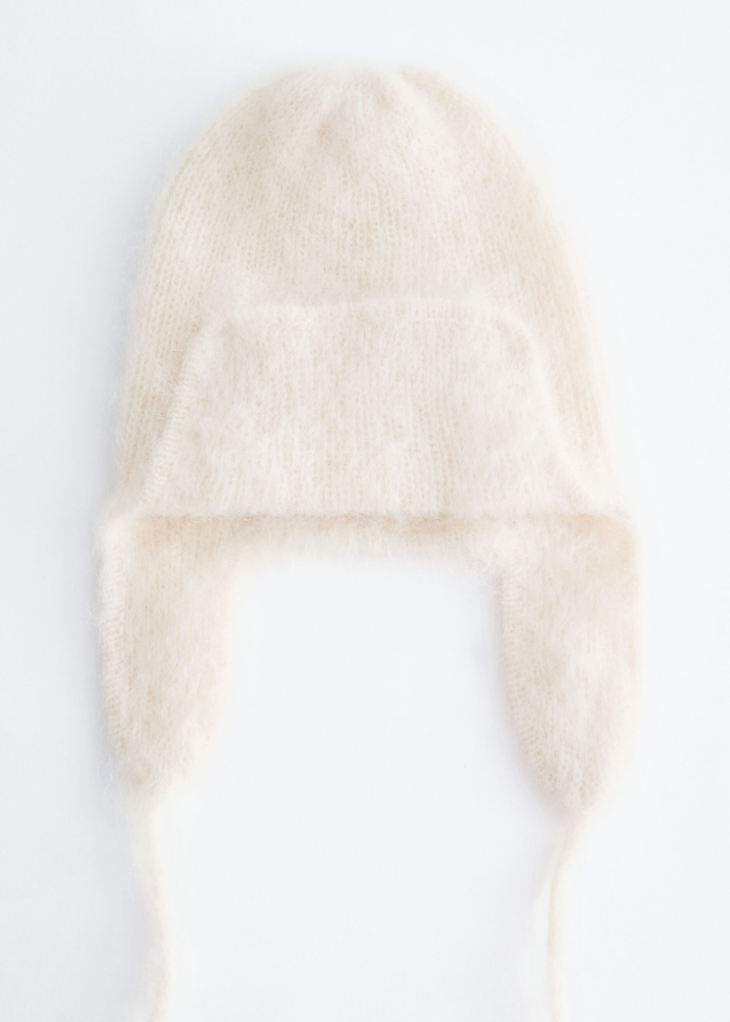 A cream-colored knit trapper hat is shown facing forward, highlighting its fuzzy, brushed texture. The design features side ear flaps with thin hanging chin straps and a folded-up front flap.