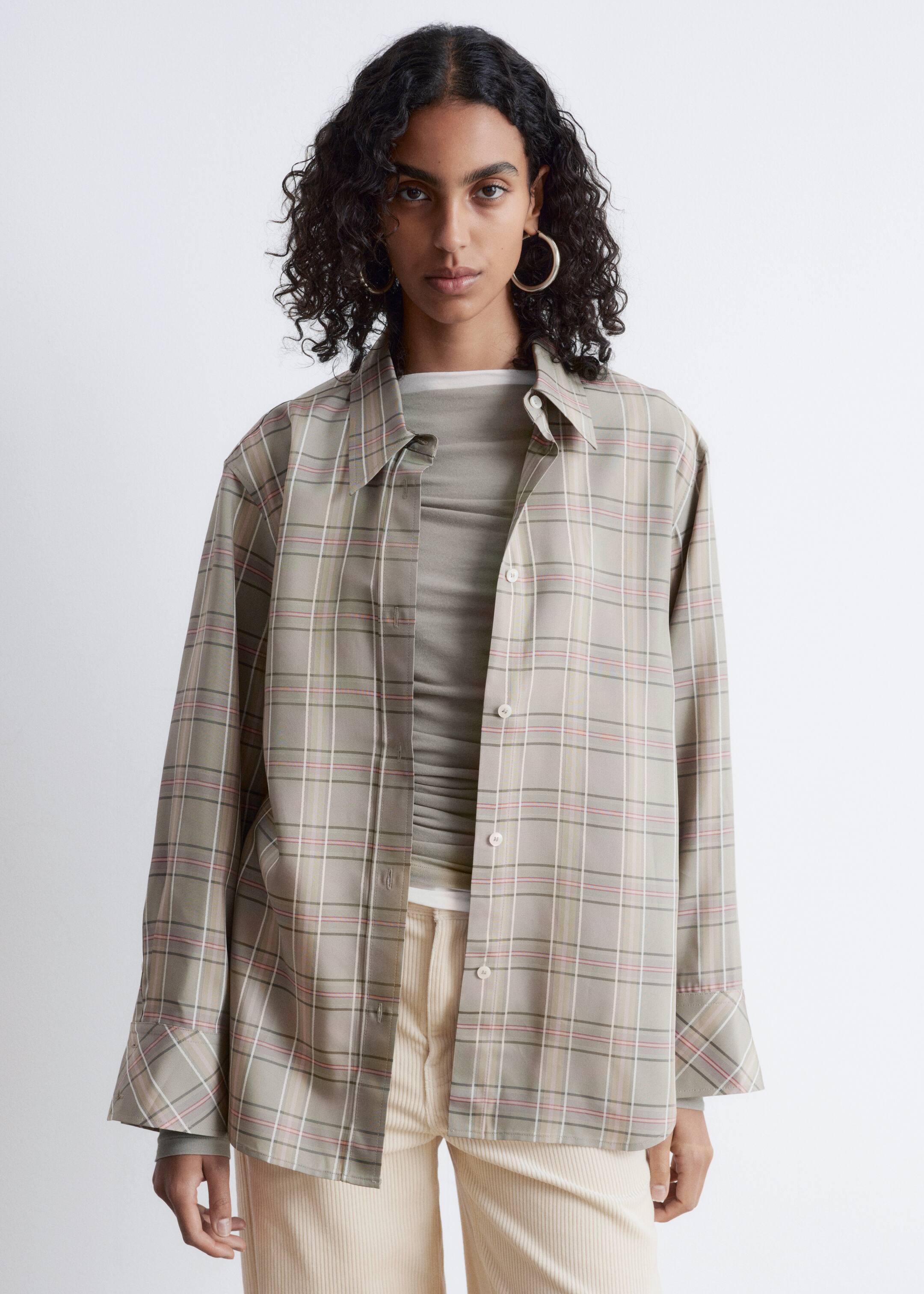 Agrandir l'image: Une chemise à carreaux aux tons neutres est présentée de face, portée ouverte sur un haut froncé à manches longues vert olive. Elle présente un col pointu, une patte de boutonnage à l'avant avec des boutons clairs et des manches larges à poignets, avec un motif à carreaux de lignes blanches, rose clair et vert-brun foncé.
