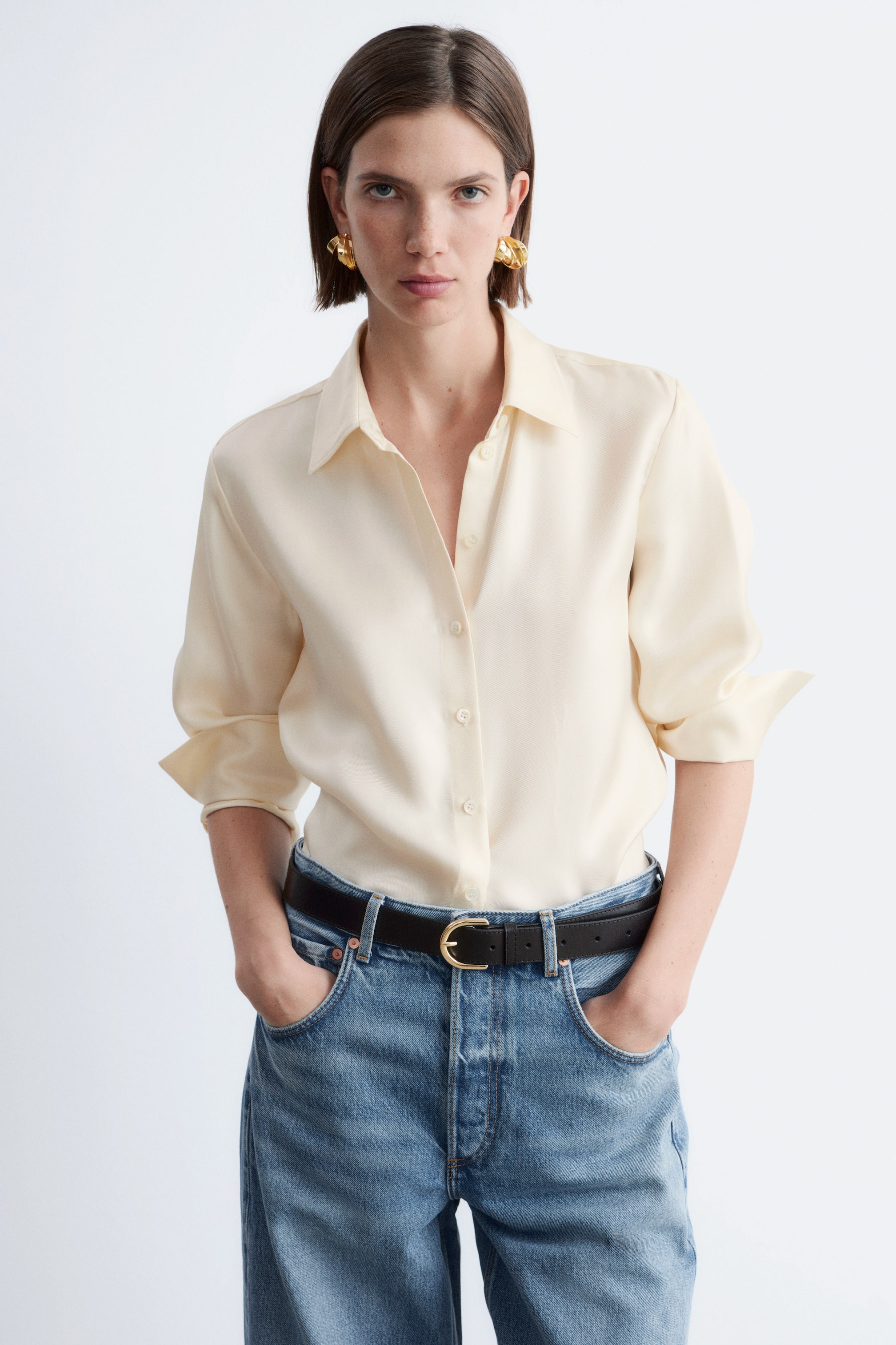 Ingrandisci l'immagine: Una camicia color crema con bottoni frontali è rivolta in avanti, caratterizzata da un colletto a punta e un'abbottonatura completa. Le maniche lunghe appaiono arrotolate fino ai gomiti, mettendo in risalto un tessuto liscio e fluente.