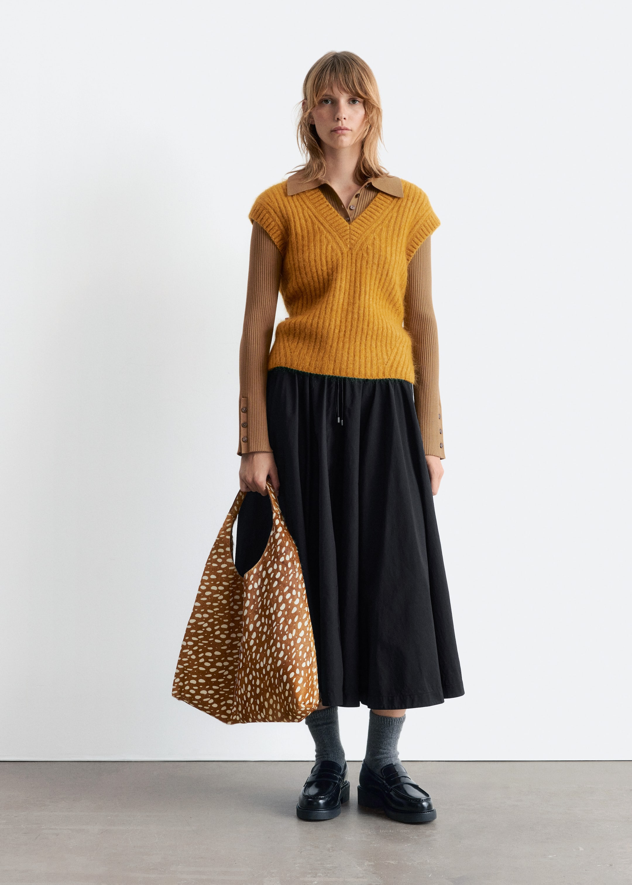 Agrandir l'image: Un gilet sans manches à col en V côtelé de couleur jaune moutarde est superposé à un polo à manches longues marron clair avec un col à revers et des poignets boutonnés. Il est assorti d'une jupe midi noire fluide, de mocassins noirs épais et de chaussettes grises, tandis qu'un sac fourre-tout marron avec un imprimé faon blanc est tenu.