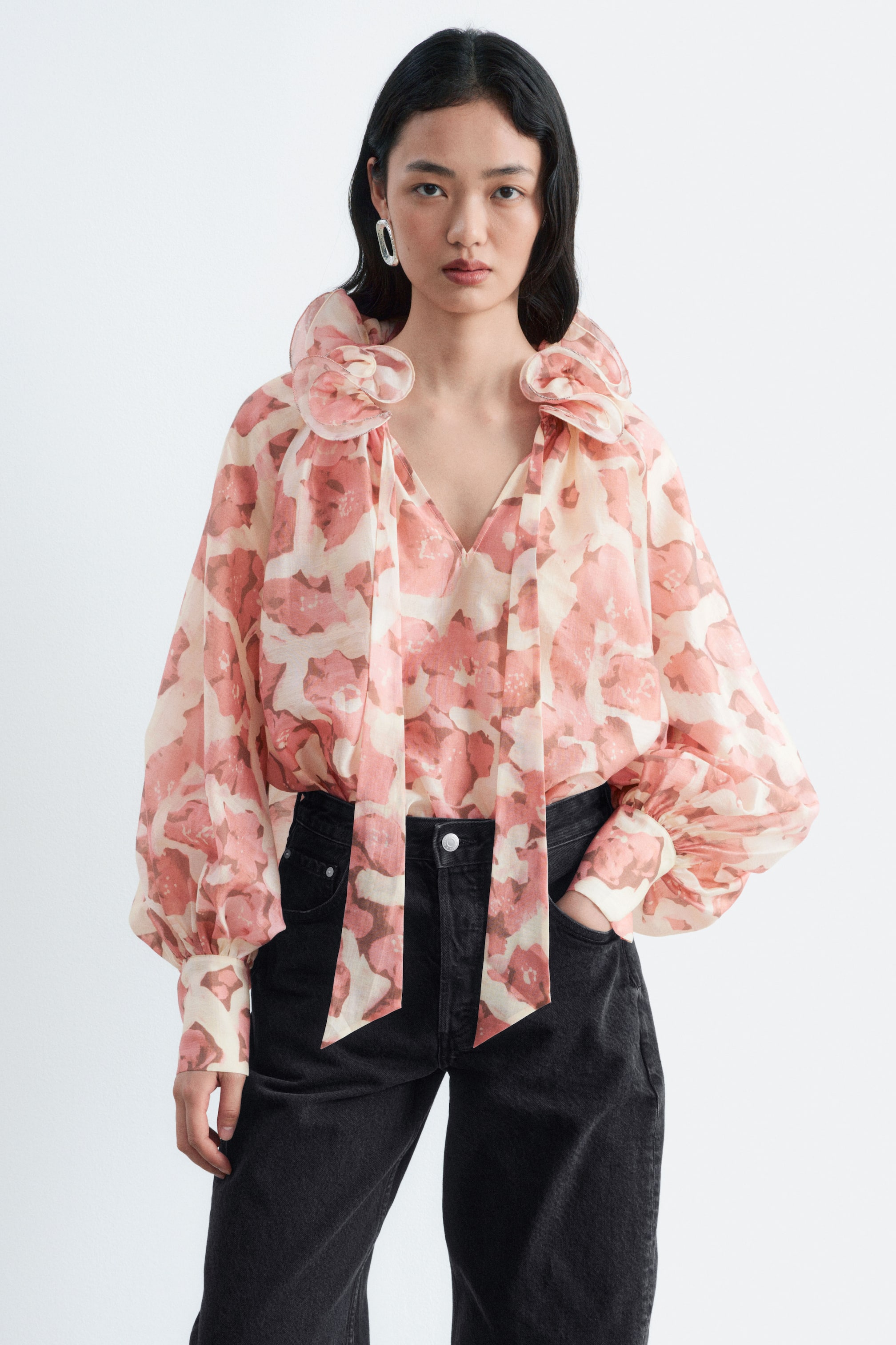 Größeres Bild anzeigen: Die Ruffle Neck Voluminous Blouse mit einem abstrakten Blumenmuster in Rosa und Creme wird vom Model getragen. Die Bluse ist mit einem auffälligen Rüschenkragen, einem V-Ausschnitt mit langen, herabhängenden Bindebändern und voluminösen Ballonärmeln gestaltet und in eine hoch taillierte schwarze Jeans gesteckt.