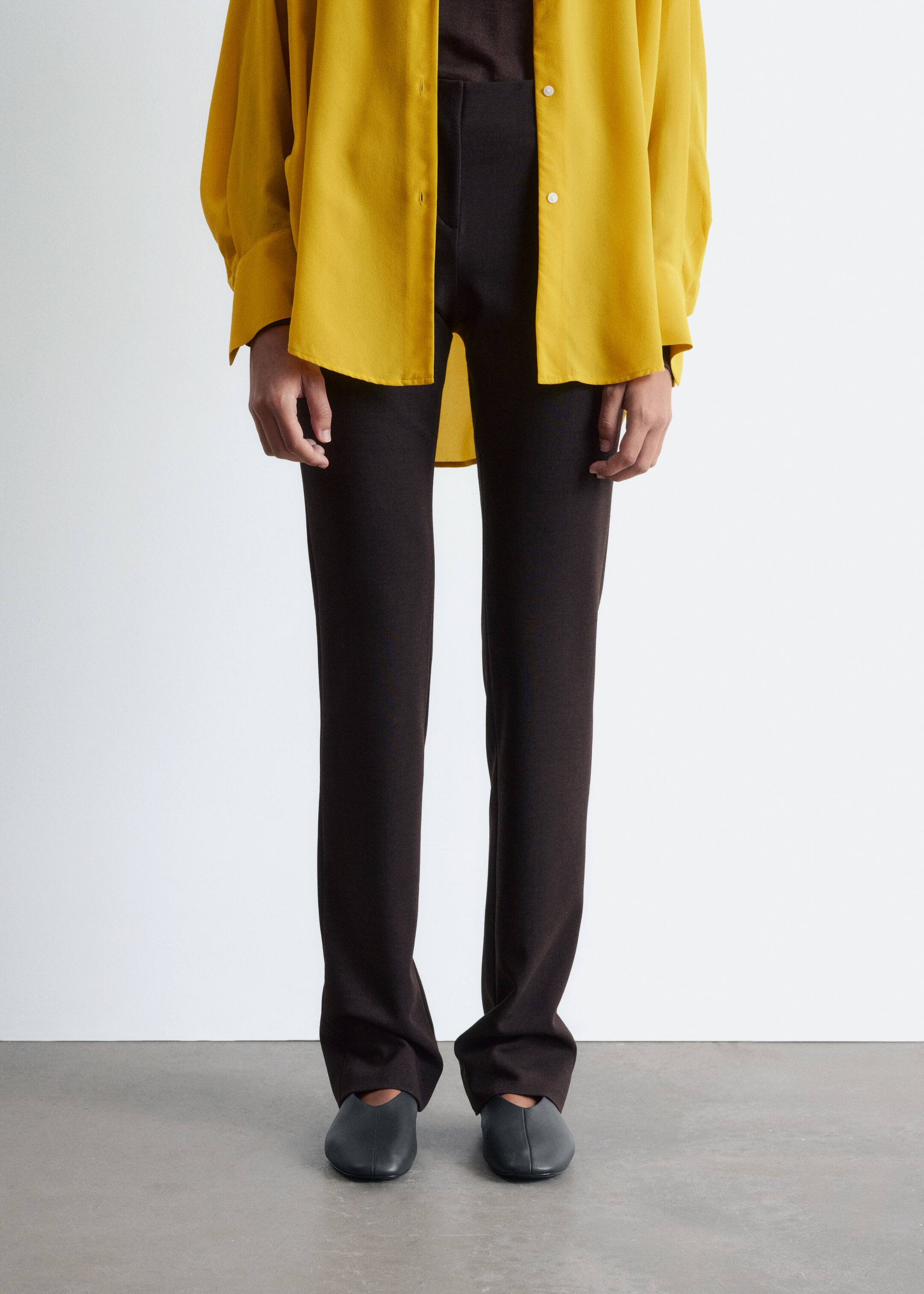Agrandir l'image: Un pantalon marron foncé est présenté de face, avec une silhouette ajustée à travers la jambe. Ils sont de longueur complète, tombant légèrement sur des chaussures plates gris foncé, et une chemise jaune surdimensionnée est superposée à leur taille haute.