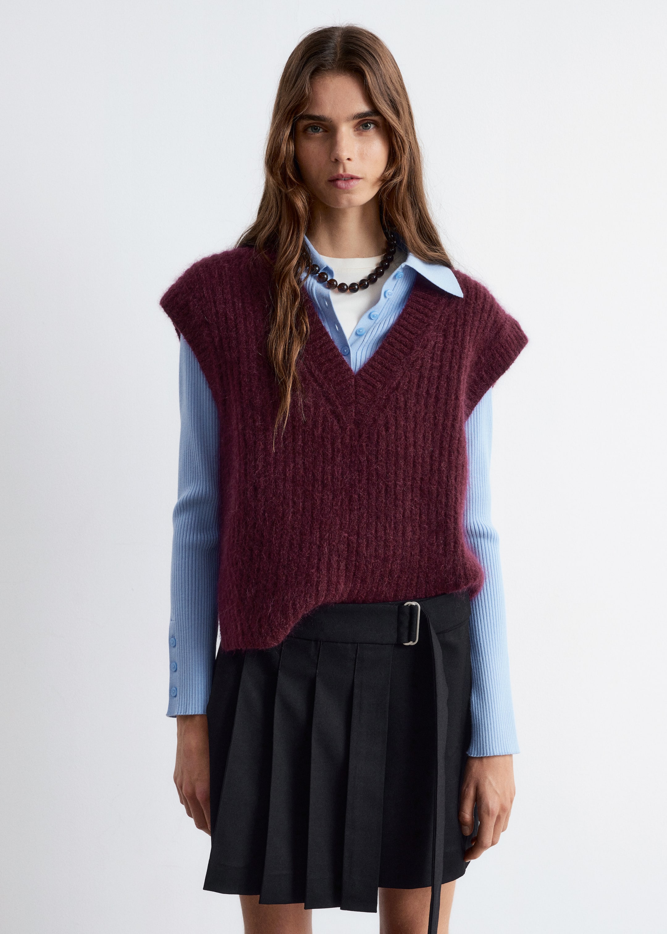Visualizza immagine più grande: Un gilet rosso scuro a V è indossato, mostrando una texture a maglia a coste e apparendo peloso, stratificato su una camicia con colletto azzurro.