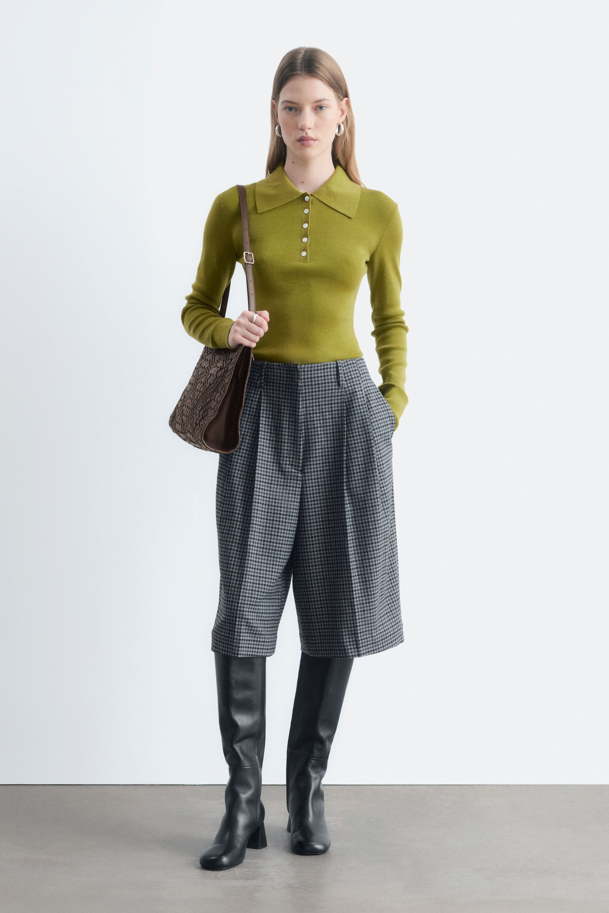 Größeres Bild anzeigen: Das Model trägt ein olivgrünes Strick-Poloshirt mit langen Ärmeln. Es verfügt über einen spitzen Kragen und eine Drei-Knopf-Leiste und ist in eine grau gemusterte Culotte gesteckt.