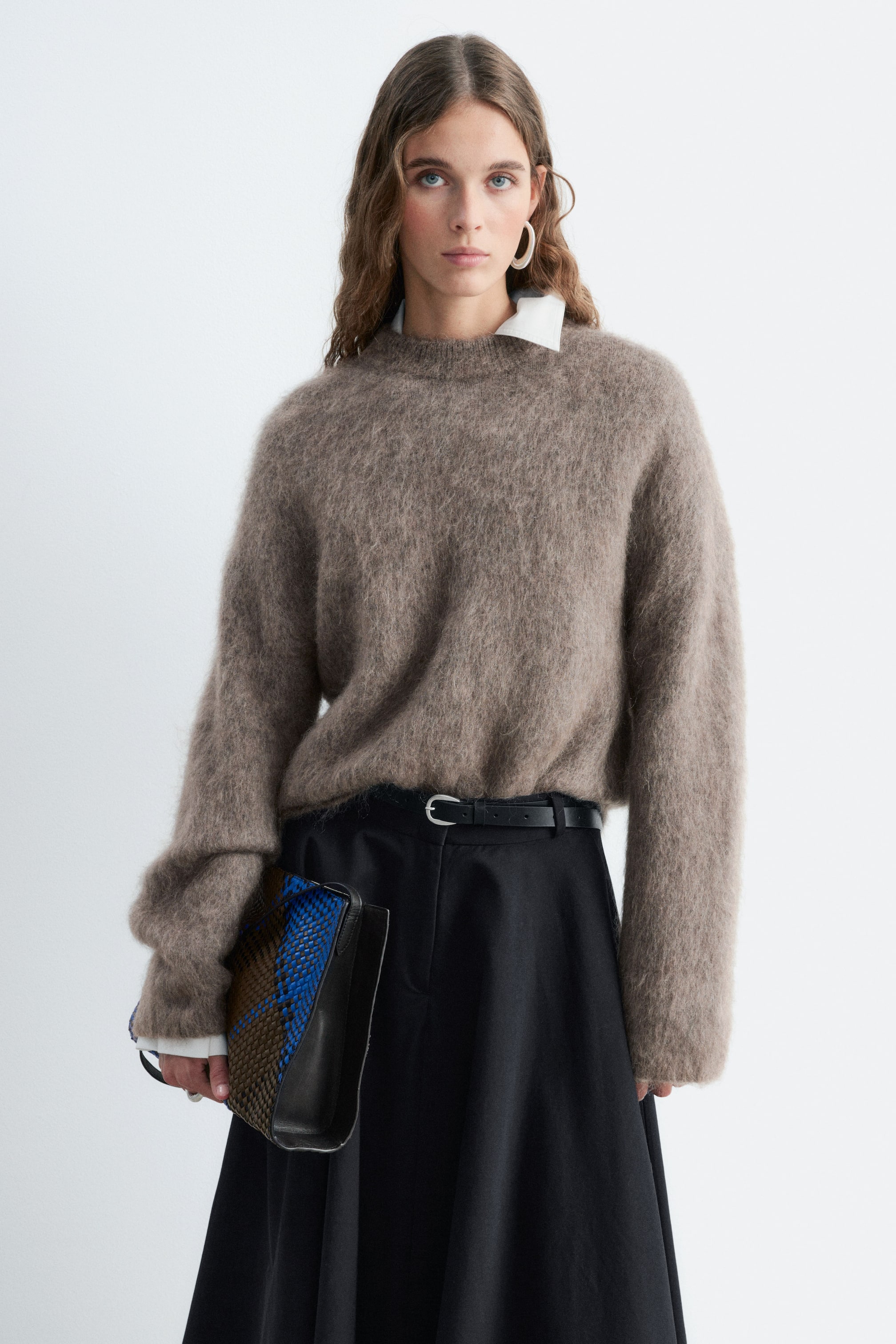 View larger image: A taupe mohair-blend jumper with a fuzzy texture and crew neck is being worn by the model, layered over a white collared shirt. The long-sleeved jumper is tucked into a black belted skirt, while the model holds a woven blue and brown clutch bag.