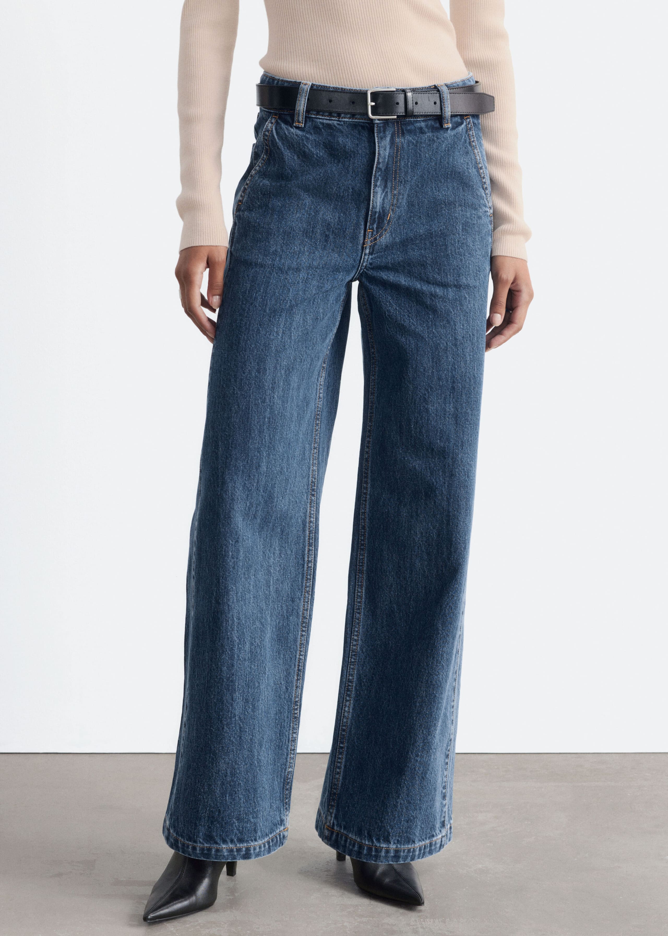 The model is wearing high-waisted, wide-leg blue denim jeans styled with a black leather belt and a tucked-in beige ribbed top. The jeans feature a medium blue wash, slanted front pockets, and are paired with black pointed-toe boots.