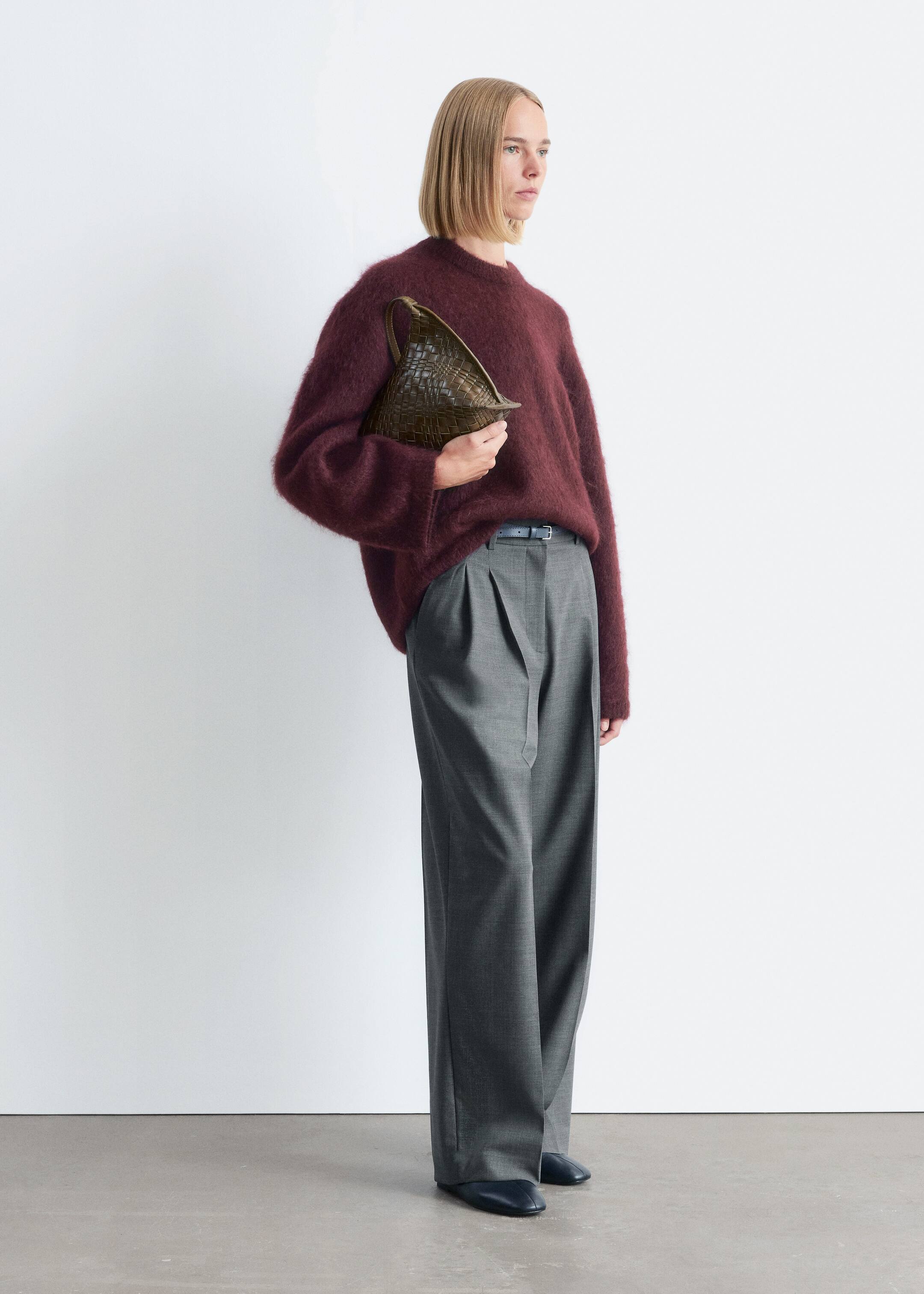 Vezi imaginea mai mare: Un model prezintă o ținută completă, având un pulover burgundy cu guler rotund, lejer și pufos, și pantaloni gri cu talie înaltă și craci largi, cu pliuri. O curea subțire și închisă la culoare este vizibilă la talie, iar o geantă triunghiulară, verde-oliv, distinctiv împletită, este ținută în mână, completând ansamblul cu pantofi negri, fără toc.