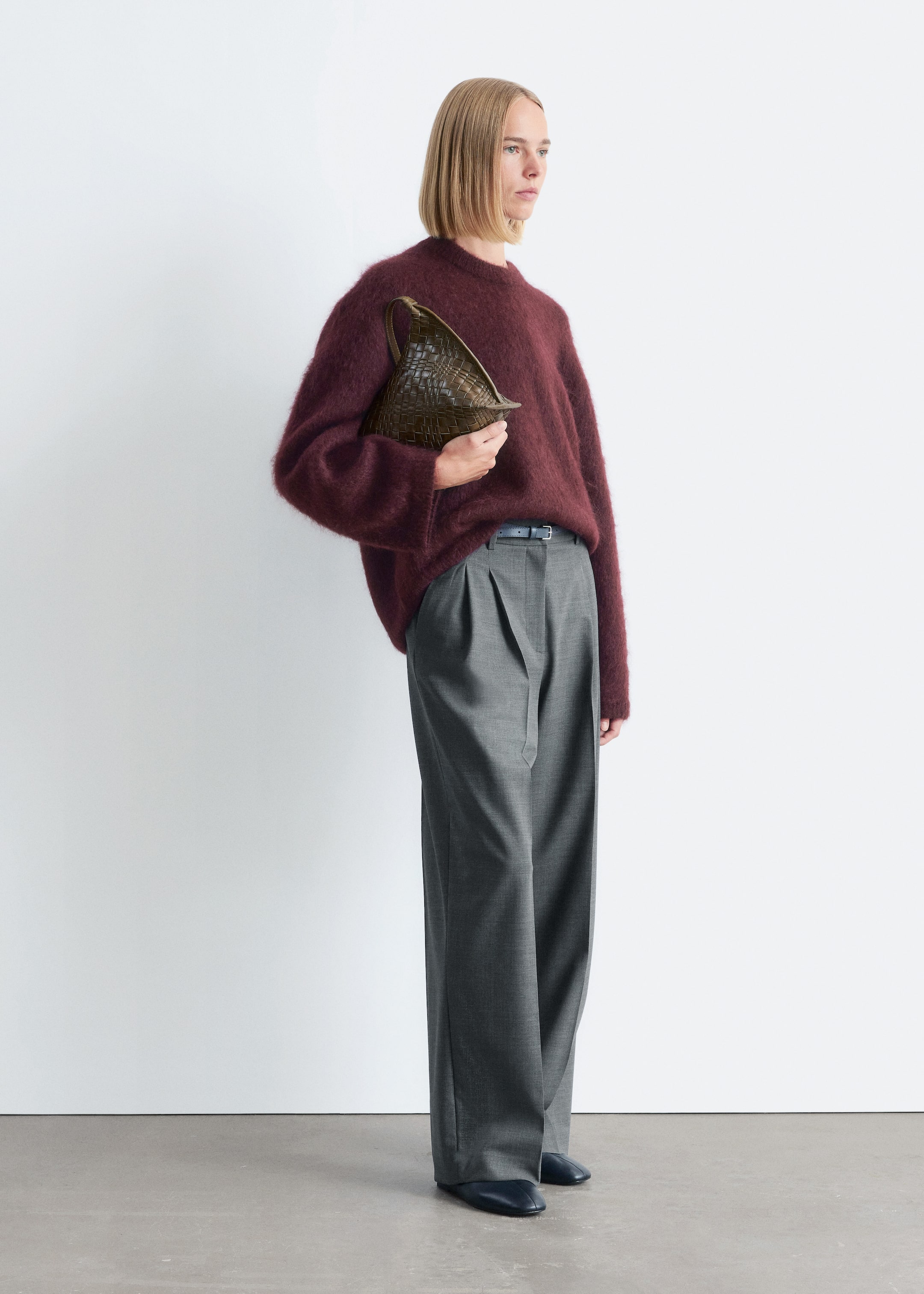 Visa större bild: En modell presenterar en hel outfit, med en löst sittande, luddig vinröd tröja med rund hals och högmidjade, vida grå plisserade byxor. Ett tunt mörkt bälte är synligt i midjan, och en distinkt vävd, olivgrön triangulär väska hålls, vilket kompletterar ensemblet med mörka, platta skor.