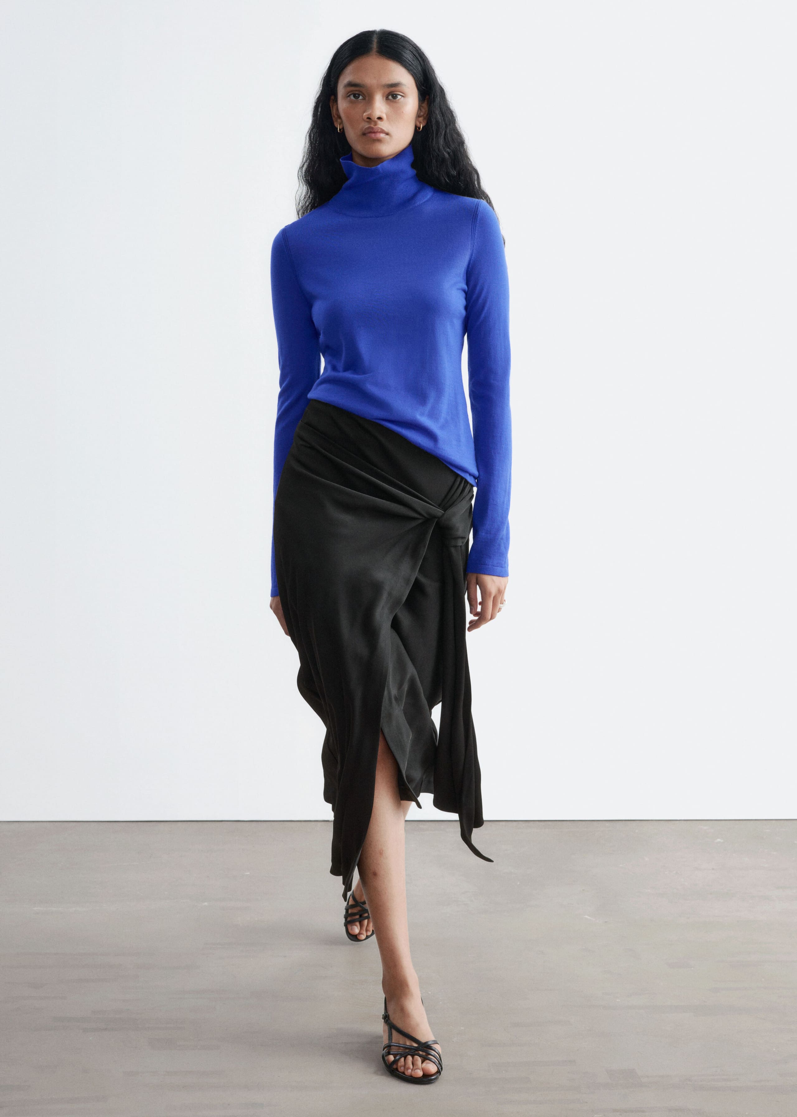 Een felblauwe polo met col van merinowol, met een hoge kraag en lange mouwen, wordt gedragen door het model. De top is gestyled met een gedrapeerde zwarte midi-rok en zwarte sandalen met bandjes terwijl het model naar voren loopt.