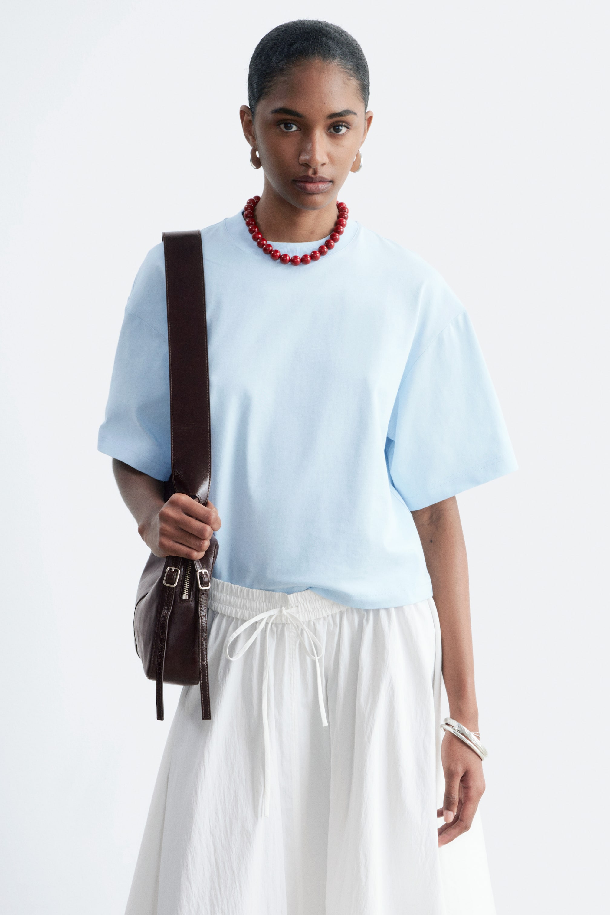 Visualizza immagine più grande: La modella indossa una t-shirt azzurra con una vestibilità rilassata e squadrata, un classico girocollo e maniche corte e ampie. La t-shirt è abbinata a pantaloni bianchi a gamba larga e una collana di perline rosse, mentre la modella porta una borsa a tracolla marrone.