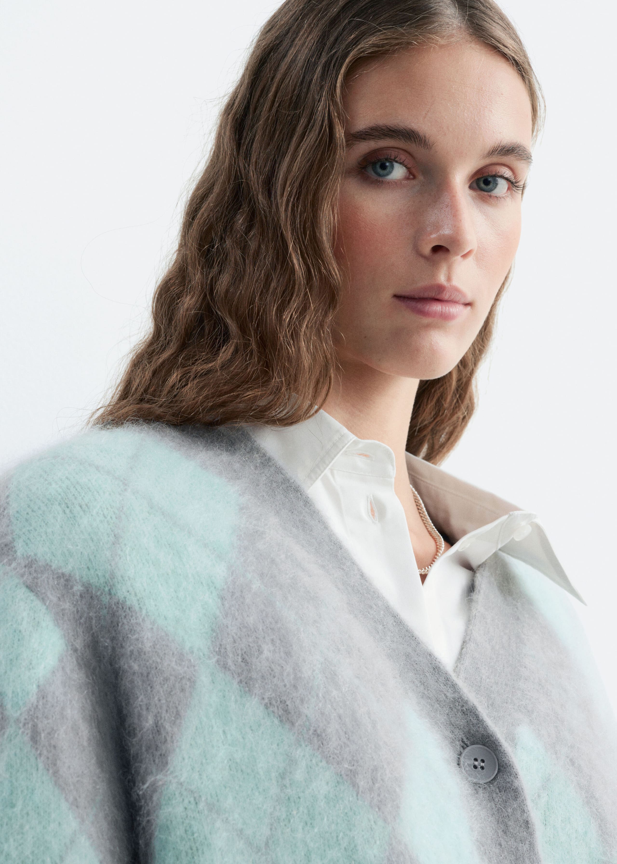 A mohair-blend argyle cardigan in gray and mint green is worn by the model. The fuzzy-textured cardigan features a V-neckline with a button closure and is layered over a white collared shirt.