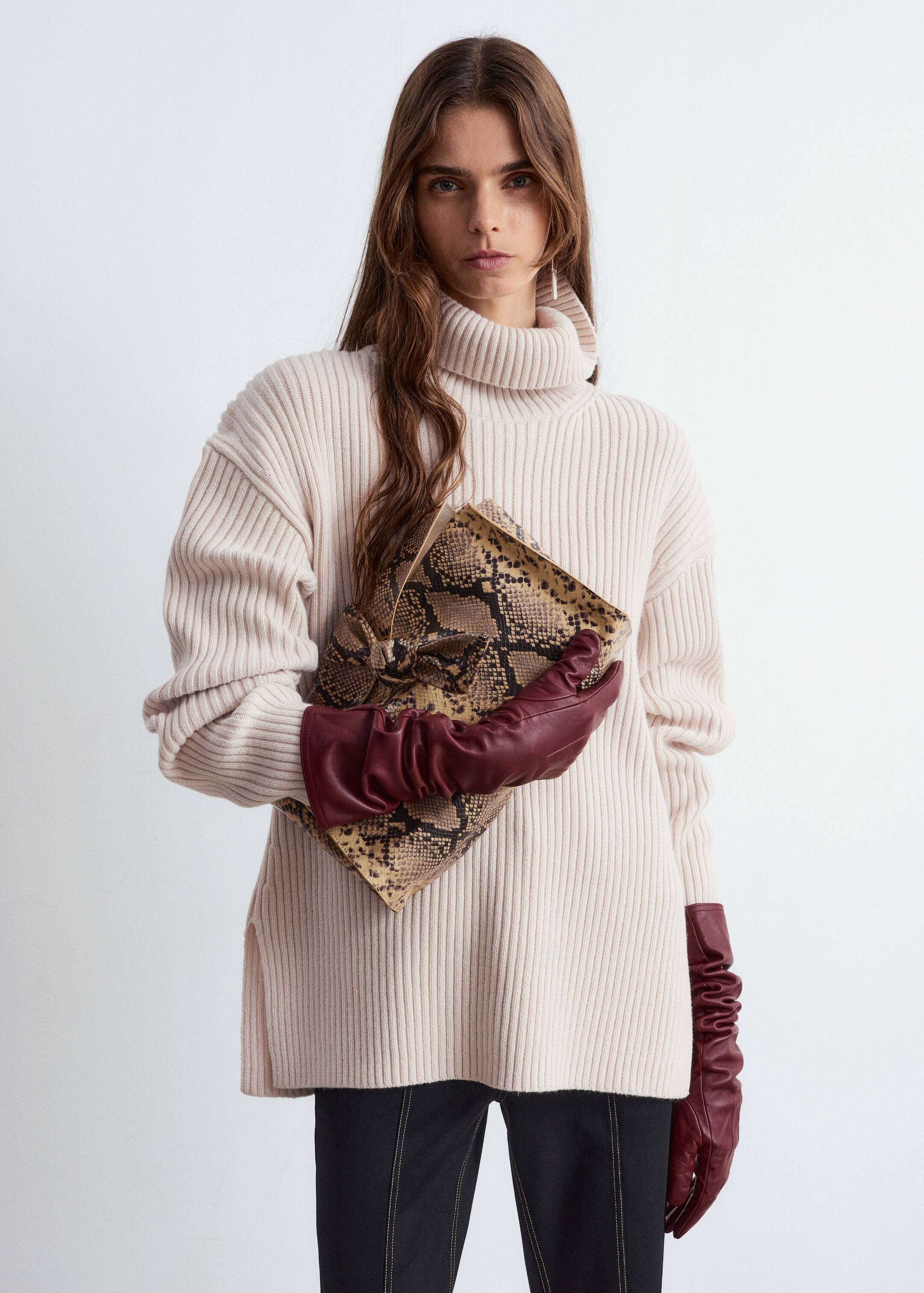 Größeres Bild ansehen: Ein cremeweißer, gerippter Strickrollkragenpullover wird von vorne präsentiert, der seine entspannte, übergroße Passform mit langen Ärmeln und breiten Manschetten zeigt. An den Säumen sind Seitenschlitze sichtbar, und eine Schlangenprint-Clutch sowie burgunderrote Lederhandschuhe werden gegen den Pullover gehalten, um seinen Fall zu demonstrieren.