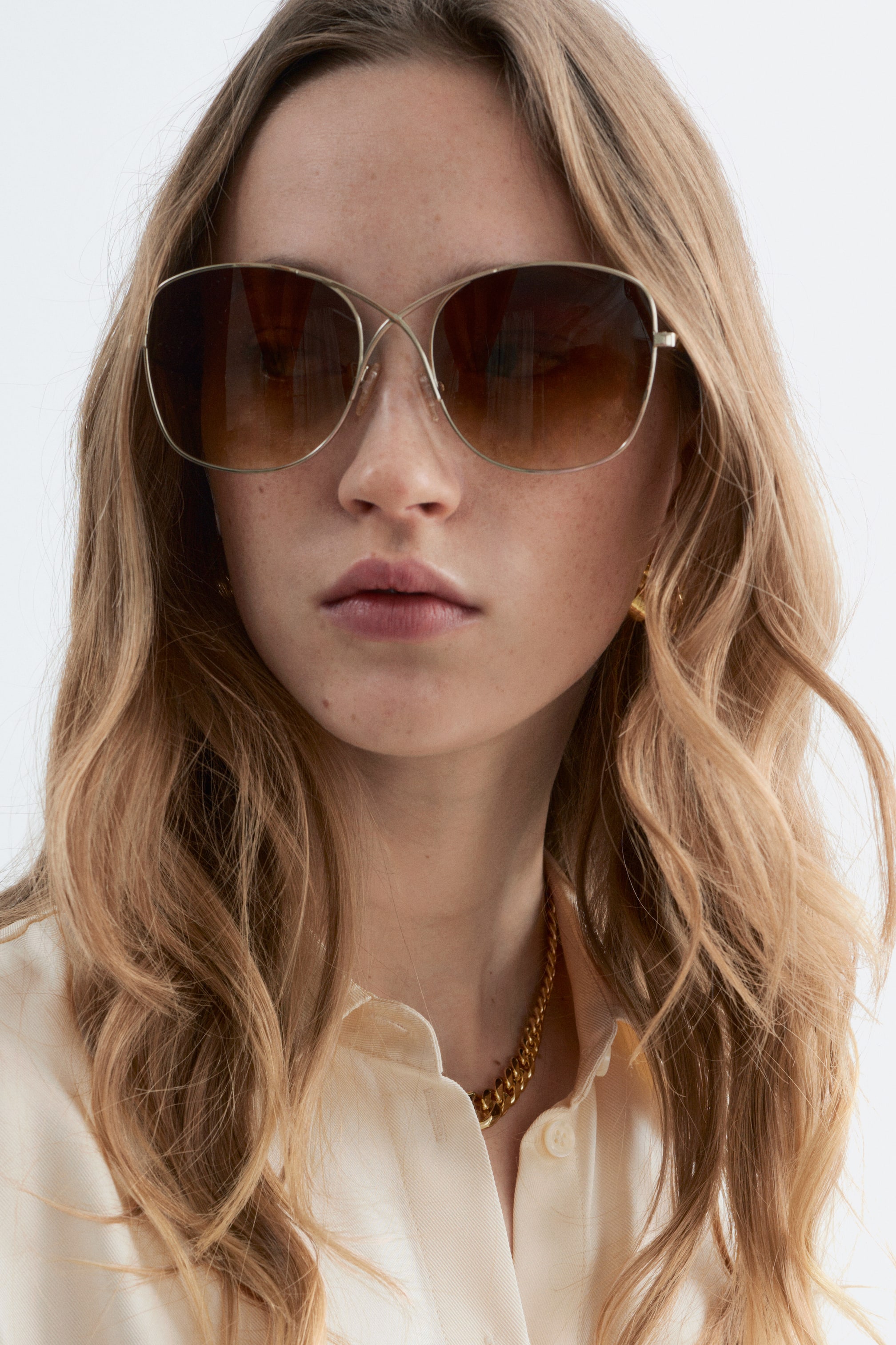 Ver imagem maior: Óculos de sol estilo aviador com tonalidade dourada e lentes grandes, ligeiramente ovais, em degradê castanho, estão a ser usados pela modelo. As armações finas cruzam-se na ponte frontal.