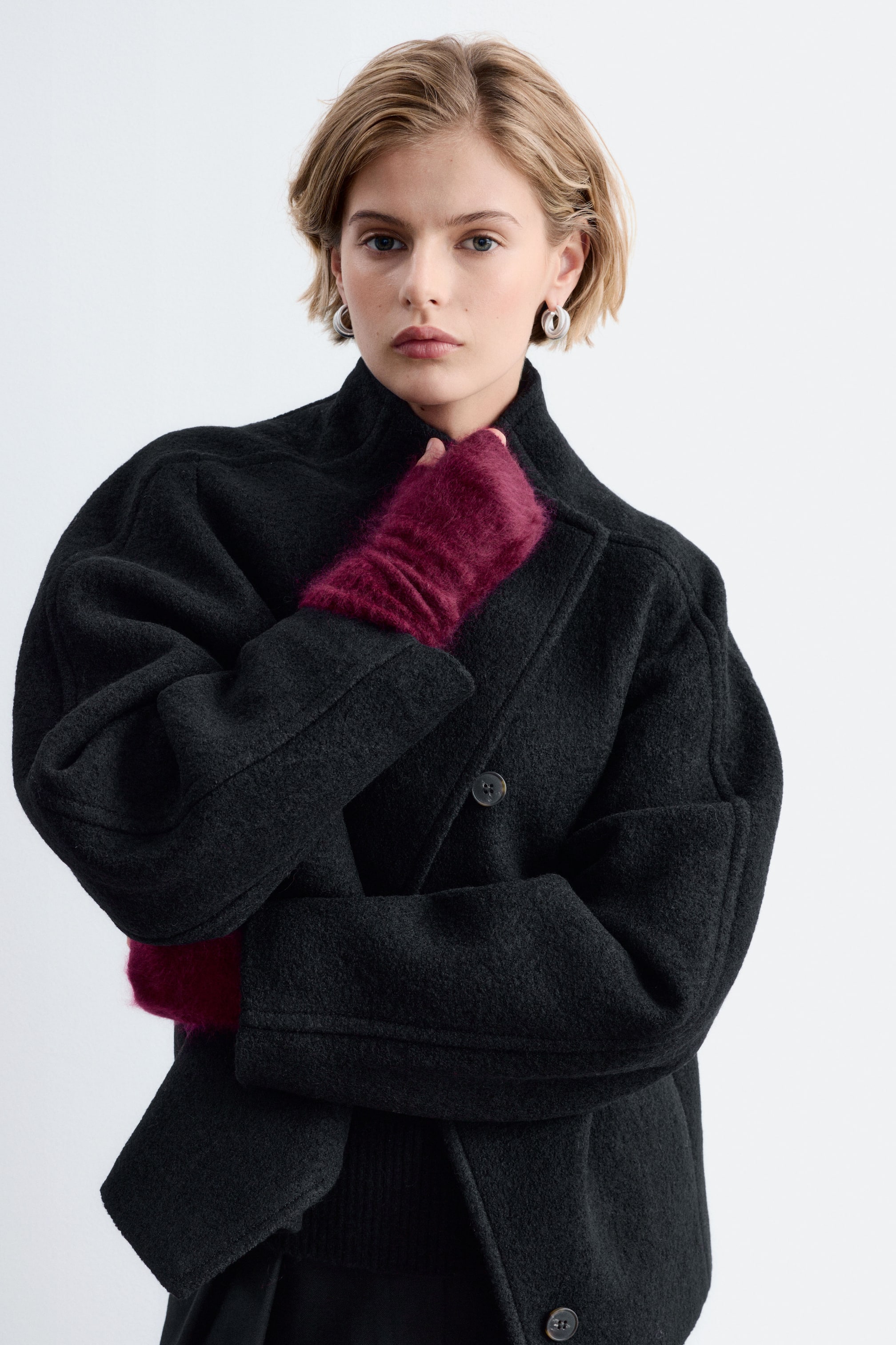View larger image: Burgundy fingerless gloves crafted from brushed cashmere are worn by the model, layered beneath the wide sleeves of a black coat. One gloved hand is raised towards the collar, highlighting the soft, fuzzy texture of the knitted accessory.