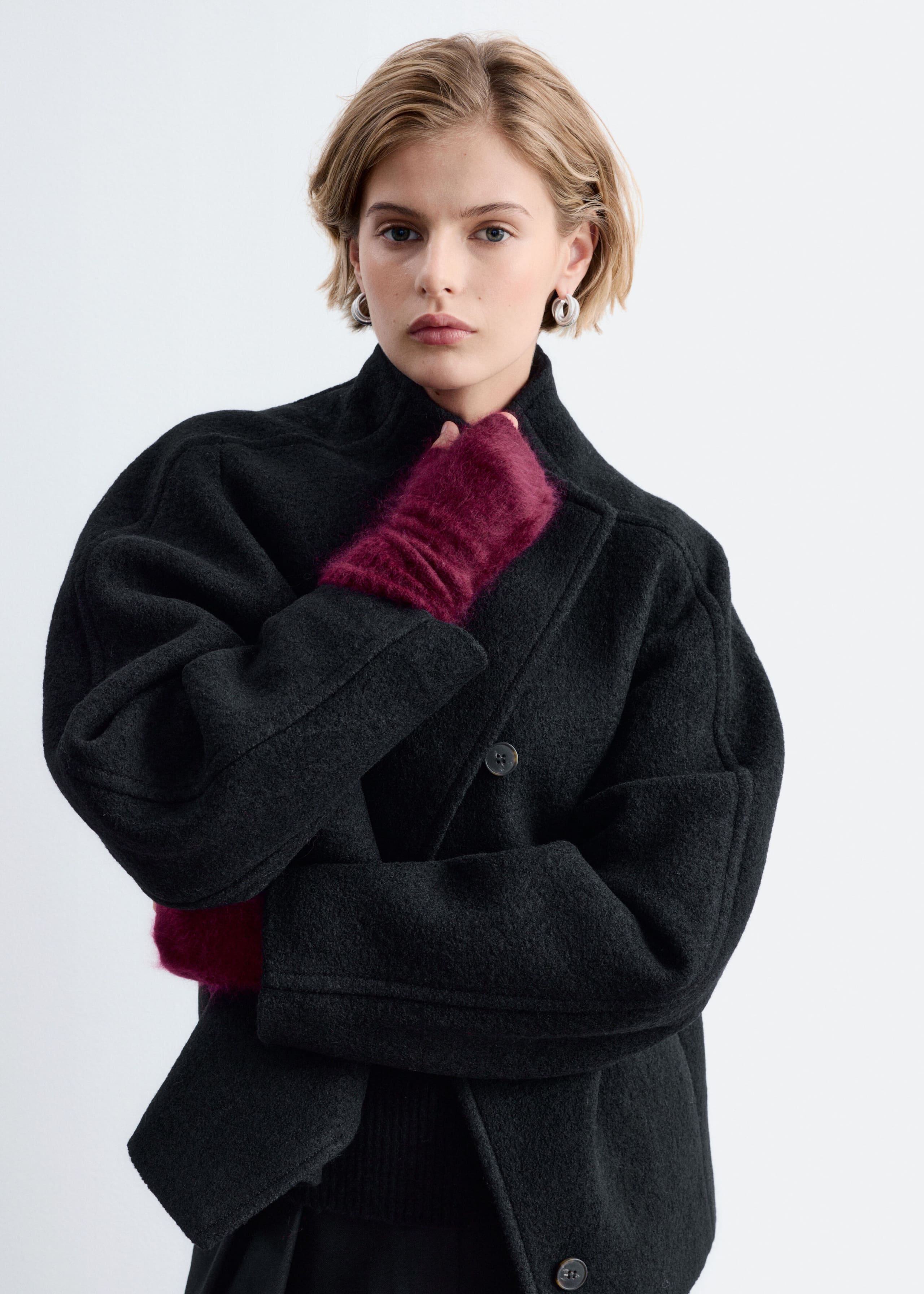 Burgundy fingerless gloves made from brushed cashmere are worn by the model, layered under the wide sleeves of a black coat. One gloved hand is raised toward the collar, highlighting the soft, fuzzy texture of the knit accessory.