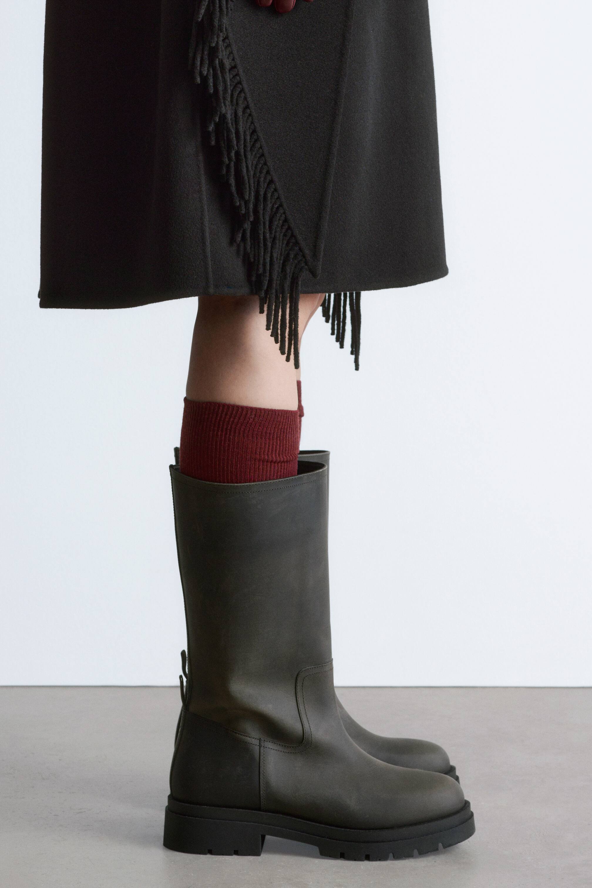 View larger image: A dark olive-green pull-on boot is presented in side profile, facing right, featuring a matt leather mid-calf shaft and a thick black lugged sole with a low block heel. It is styled with ribbed dark-red socks and a dark skirt with a fringe.