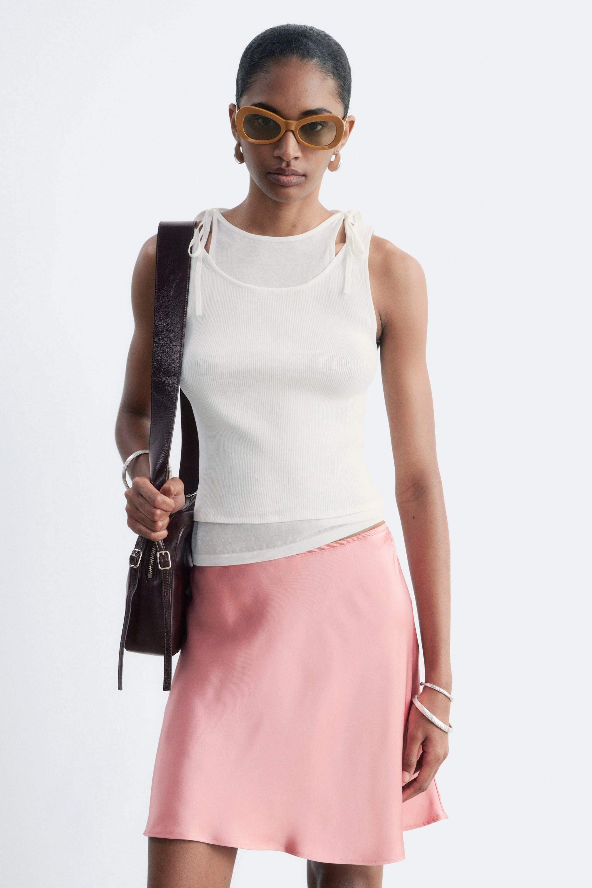 View larger image: The model is wearing a cream double-layered vest top, featuring a ribbed outer layer with a round neckline and an inner layer with a high neckline and self-tie shoulder details. A dark brown bag strap crosses the shoulder, and the vest top is paired with a pink skirt.