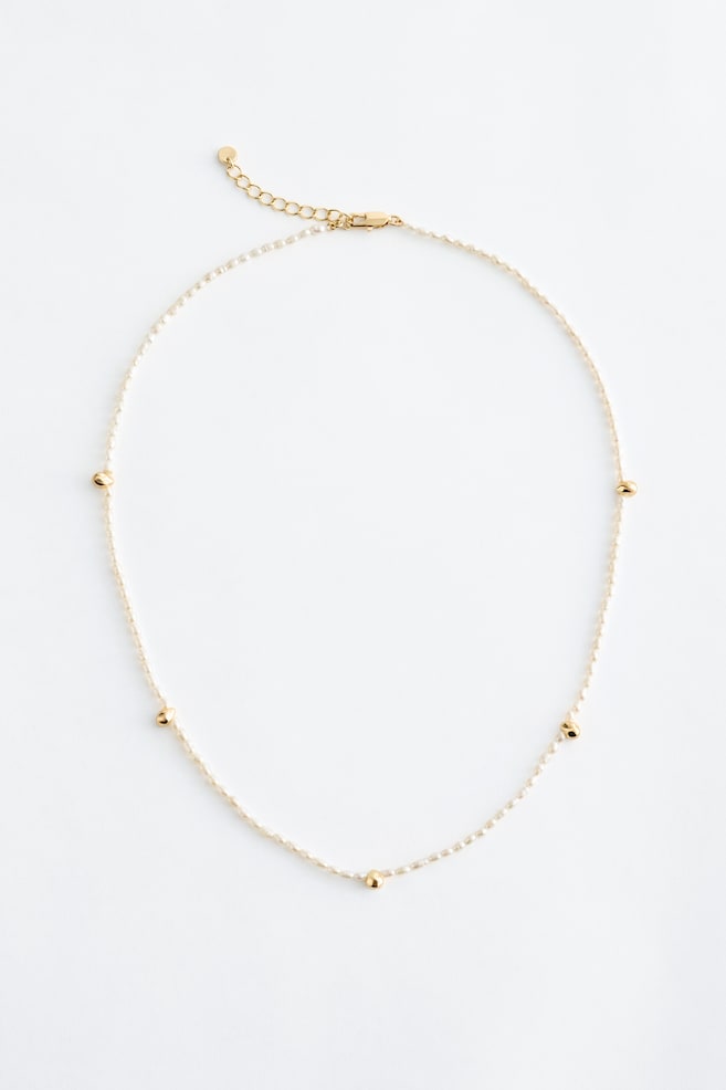 A delicate necklace featuring a chain of small, light-coloured seed beads, interspersed with five round, gold-toned beads. A gold-coloured lobster clasp and an extender chain are visible at one end. - 1