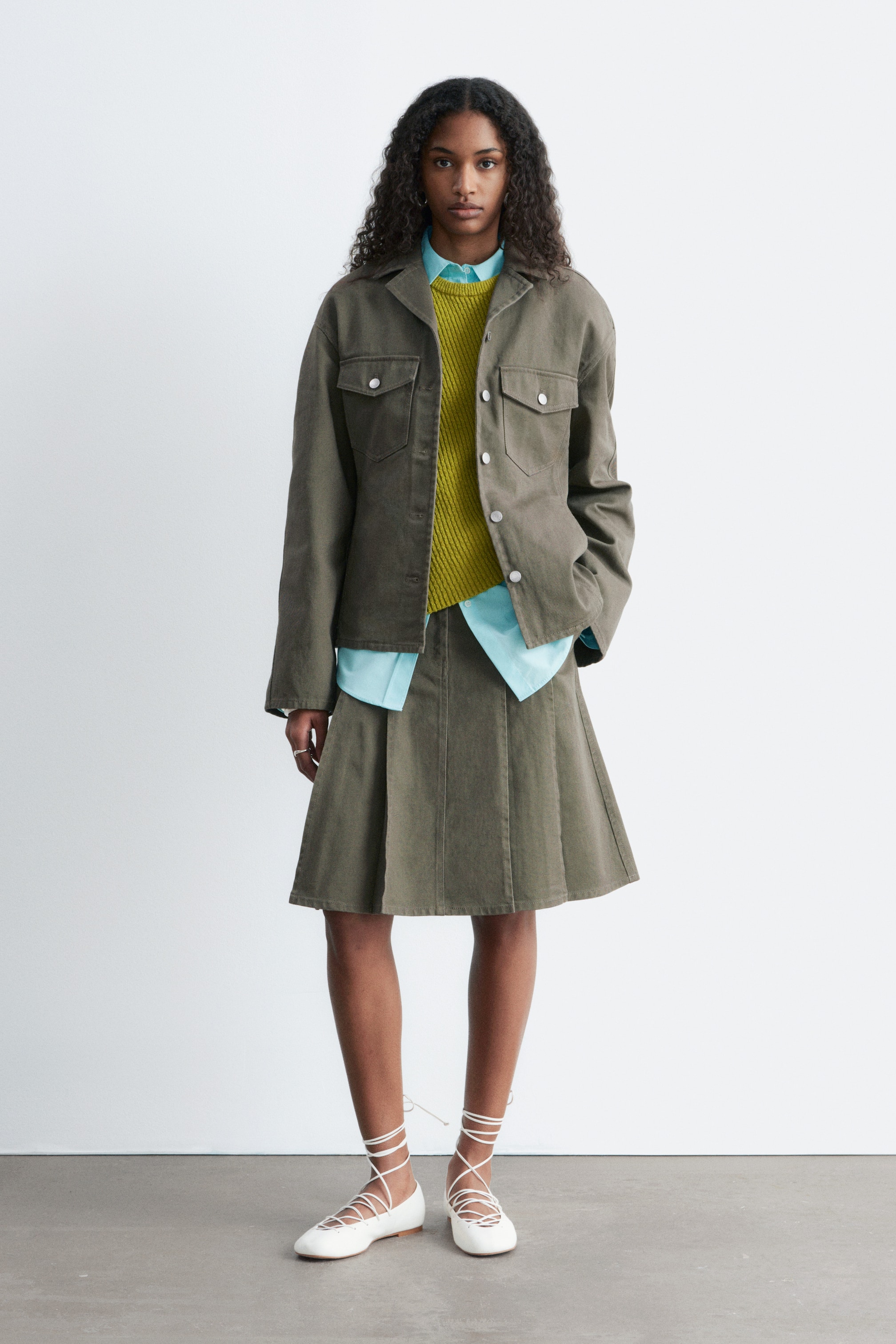 View larger image: An outfit features a dark olive utility jacket with two chest flap pockets and two side pockets, layered open over a bright lime green knitted jumper and a light blue collared shirt. A matching dark olive pleated A-line skirt complements the jacket, and white lace-up ballet flats complete the ensemble.