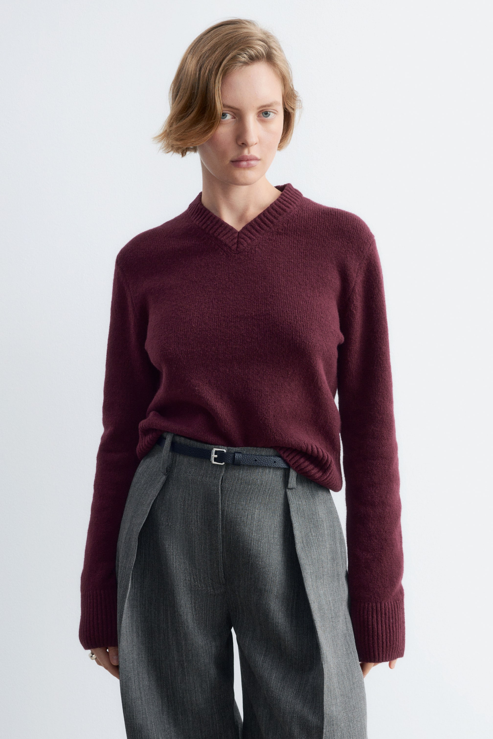 Agrandir l'image: Un pull en maille bordeaux, doté d’un col en V côtelé, de manches longues et de poignets côtelés, est porté par le mannequin. Le pull est rentré dans la taille d’un pantalon gris taille haute, ceinturé d’une fine ceinture.
