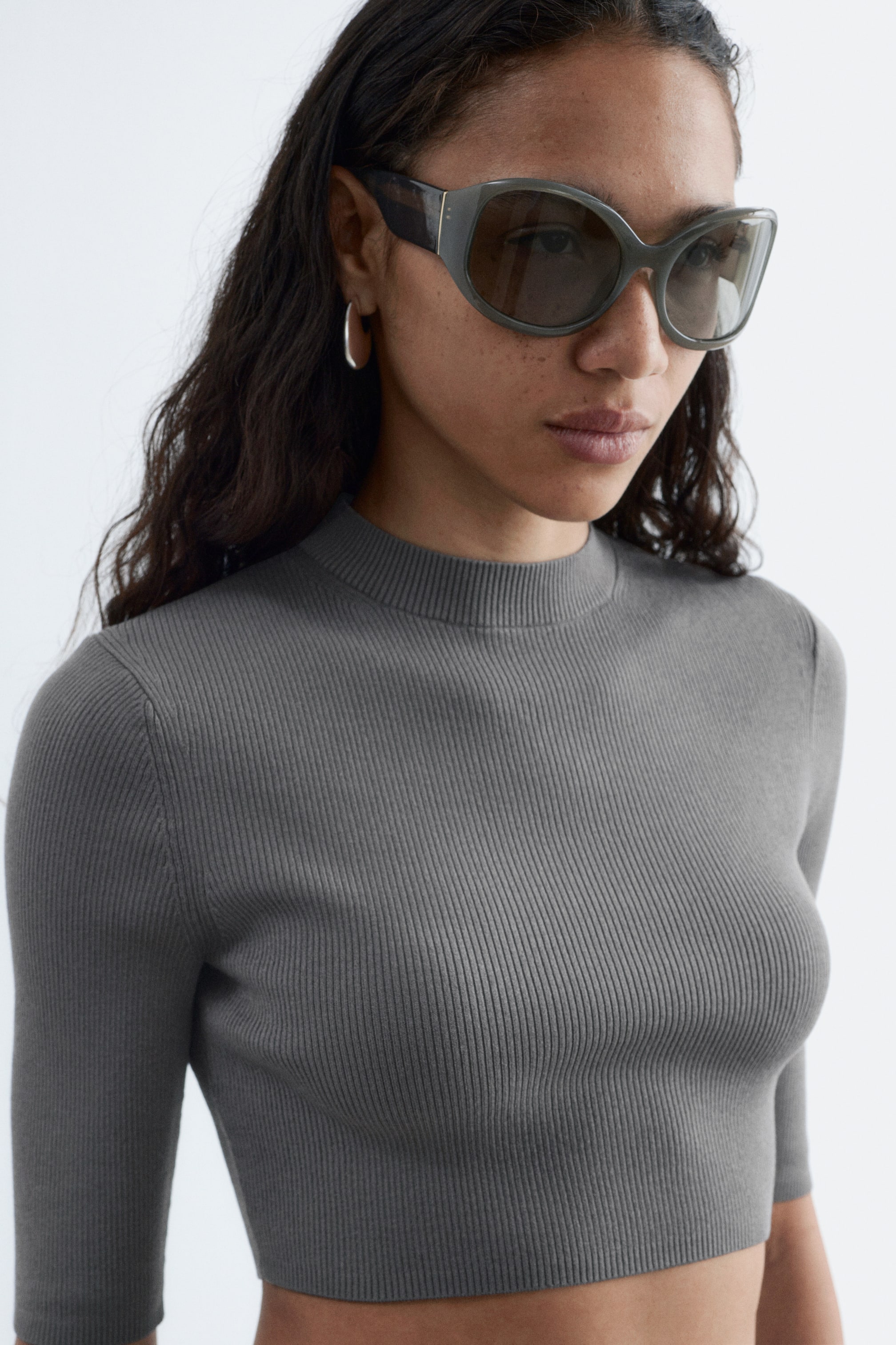 Visualizza immagine più grande: Occhiali da sole stile farfalla grigio scuro con montature spesse e lenti scure sfumate indossati, rivolti leggermente verso destra. Un top grigio aderente a coste con collo alto finto, maniche al gomito e orlo corto è indossato.