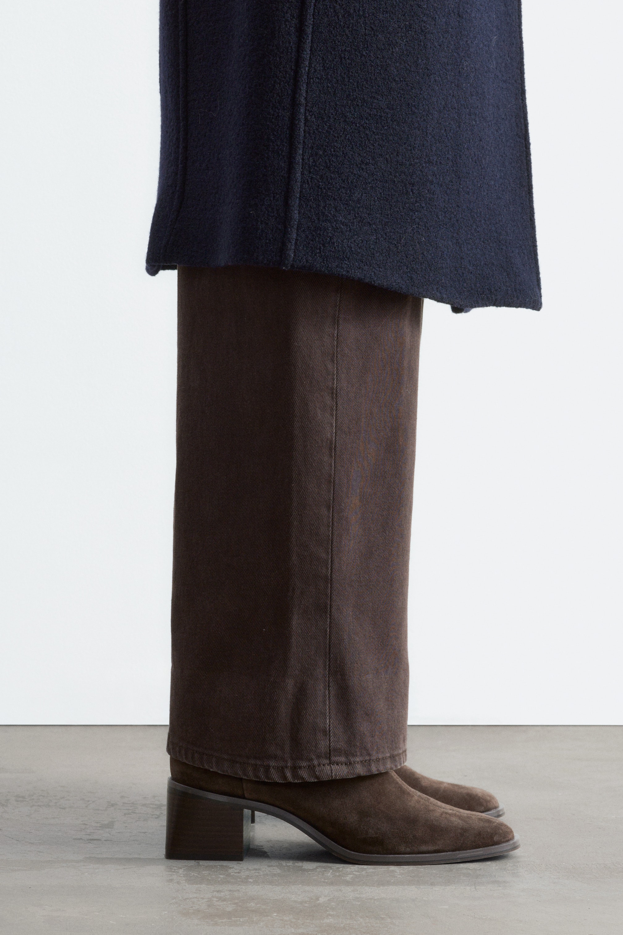 Agrandir l'image: Des bottes en daim brun foncé, à bout en amande et à talon bloc épais, sont portées par le mannequin. Les bottes sont associées à un pantalon brun à jambes larges et à un manteau bleu marine.