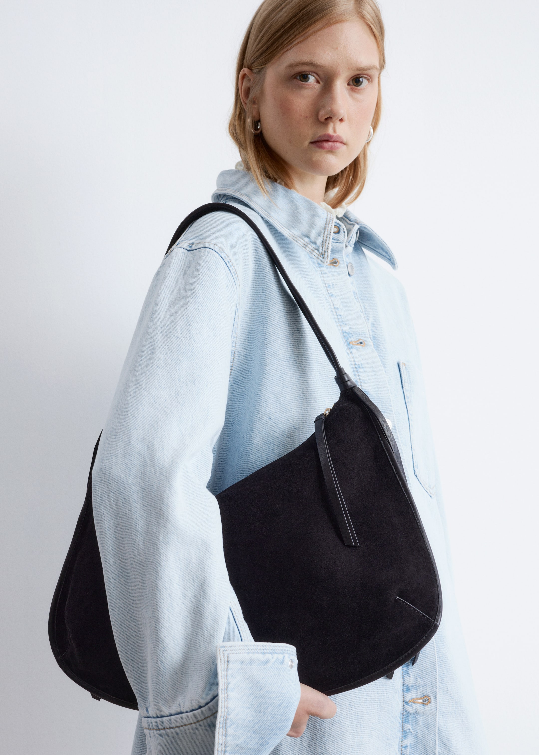 Ingrandisci l'immagine: Una borsa a tracolla in pelle scamosciata nera viene indossata, con la parte anteriore rivolta in avanti e che mostra una silhouette curva a mezzaluna. Presenta una sottile tracolla nera e una chiusura con cerniera visibile dotata di un lungo tiretto.