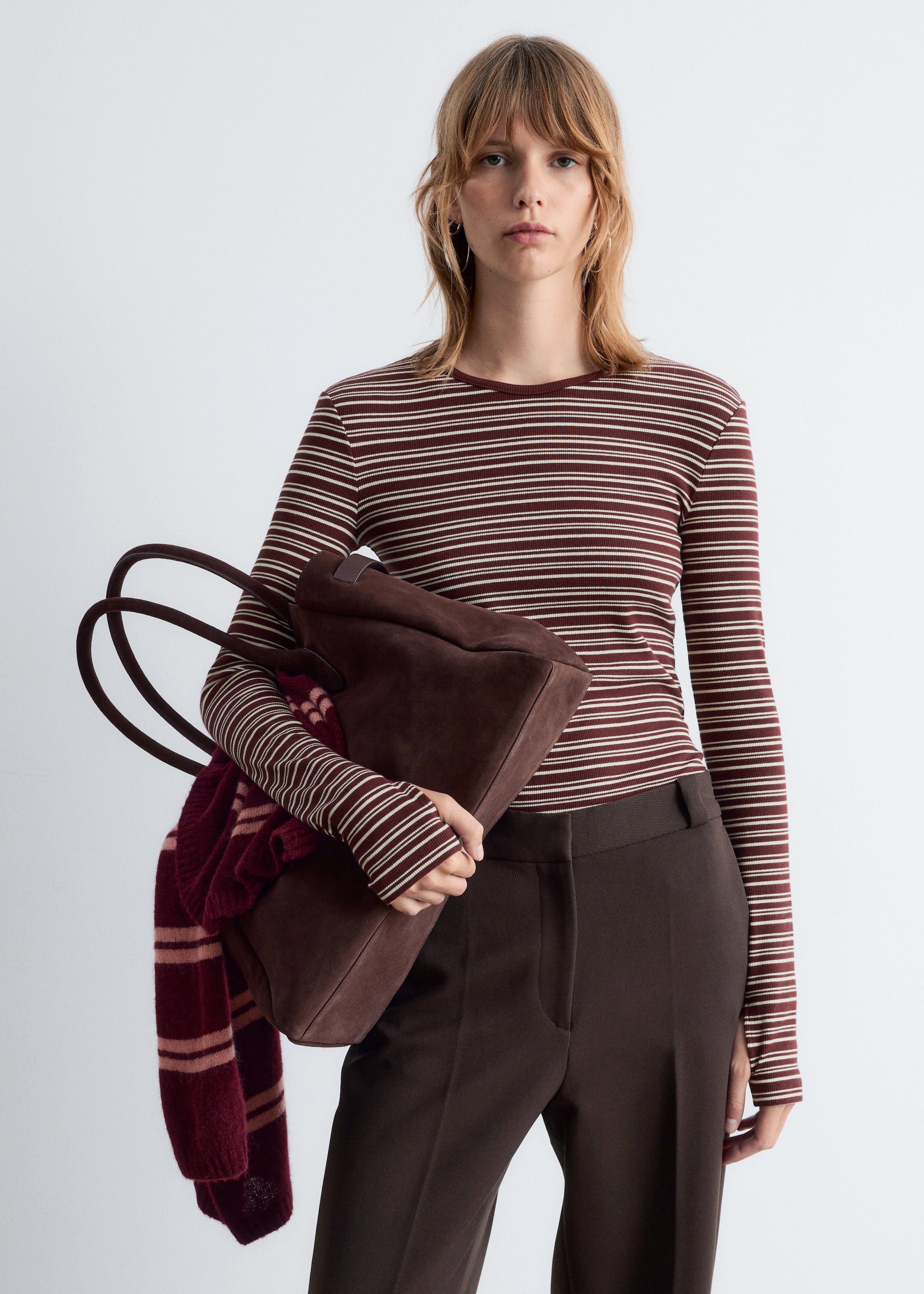 Agrandir l'image: Un haut ajusté à manches longues de couleur brun-rouge foncé avec de fines rayures horizontales blanc cassé est porté, assorti à un pantalon marron foncé à devant plat. Un sac fourre-tout en daim marron foncé avec deux poignées supérieures est tenu, d'où pend une écharpe rayée brun-rouge.