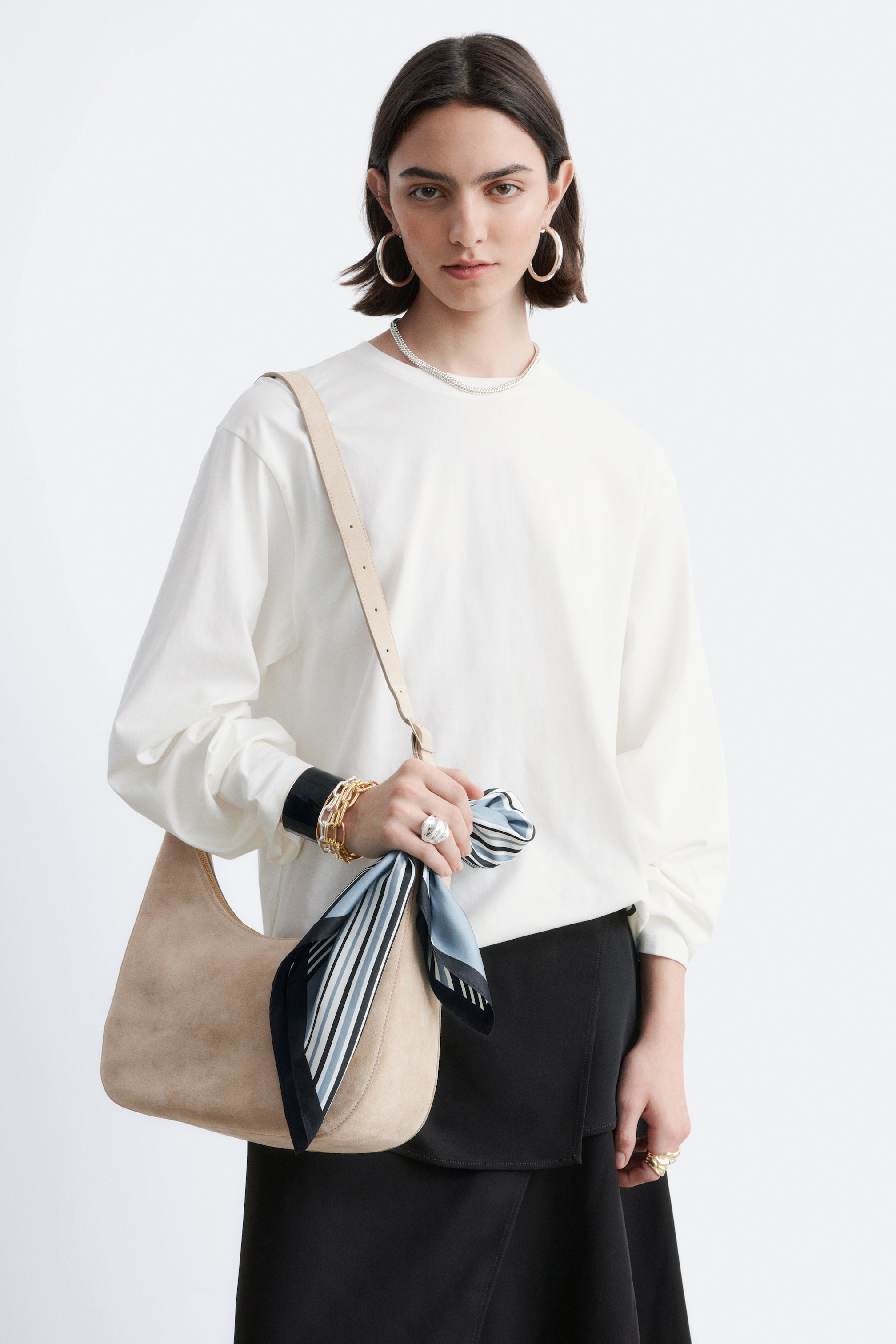 Nagyobb kép megtekintése: Egy bézs színű, velúr félhold váll táska látható elölről, amely bemutatja a puha, laza szerkezetét. Egy világos színű pánt látható a vállon, és egy fekete, fehér és világoskék csíkos sál van a táska oldalára kötve.