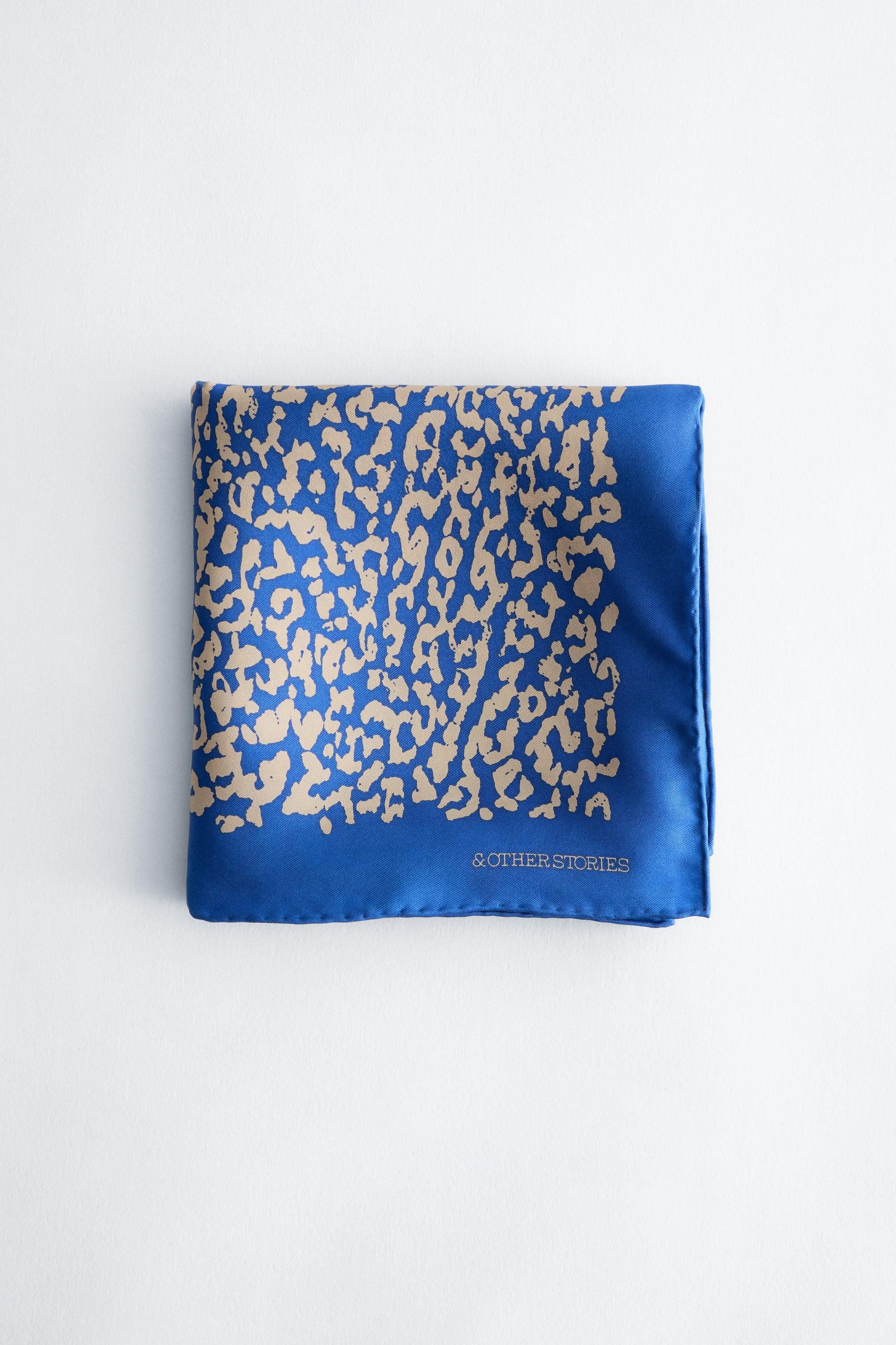 Grotere afbeelding bekijken: Een bedrukte zijden sjaal, vierkant gevouwen en van voren weergegeven, met een levendige blauwe basis en een beige abstract dierenprintpatroon. De sjaal heeft een effen blauwe rand langs de rechter- en onderrand, met de merknaam "& OTHER STORIES" in kleine goudkleurige letters gedrukt.