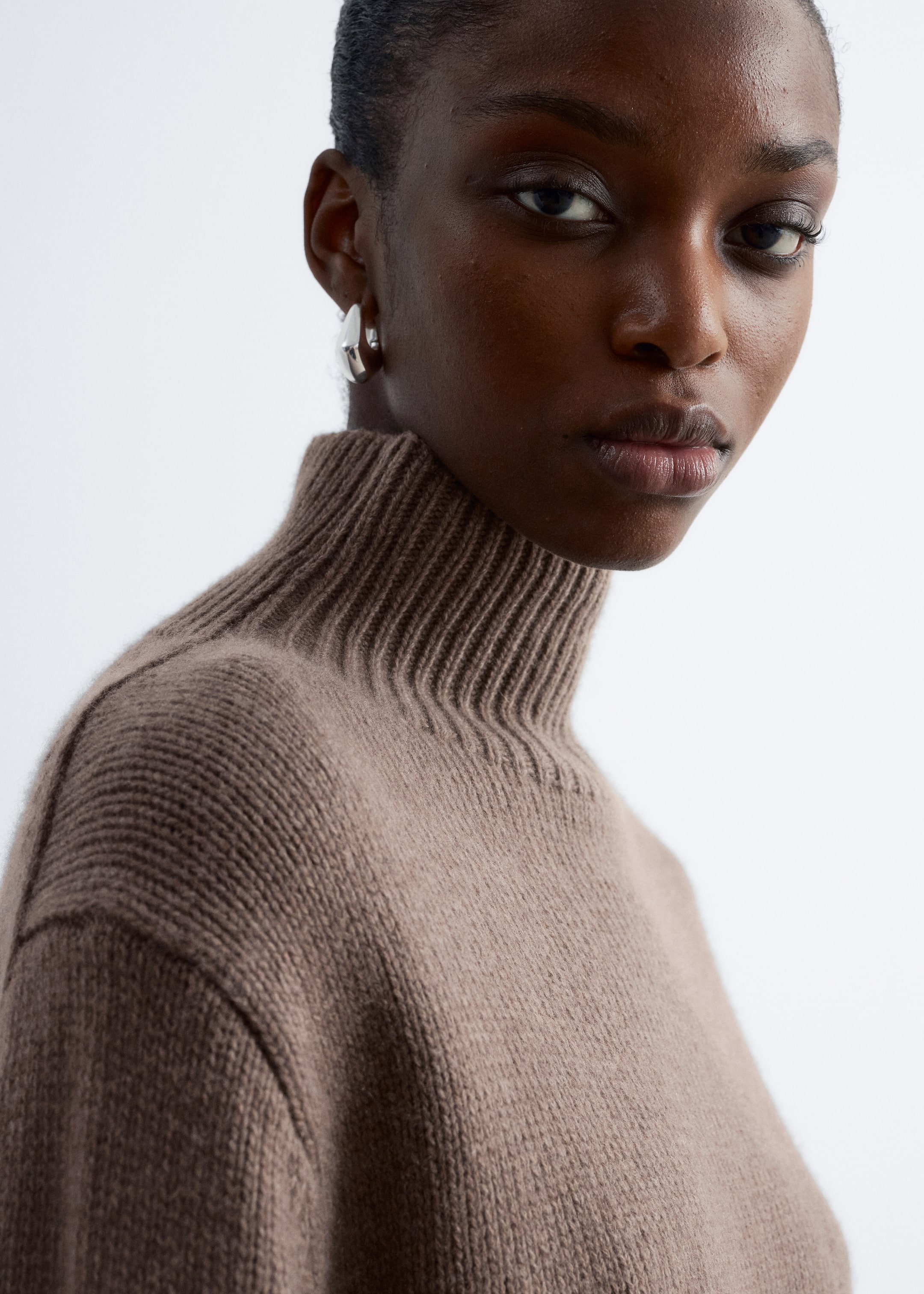 Grösseres Bild ansehen: Ein brauner Rollkragenpullover wird getragen, der einen gerippten Rollkragen und einen glatt gestrickten Körper aufweist. Der Pullover ist zur Seite geneigt, was seine lockere Passform um die Schulter herum zeigt.