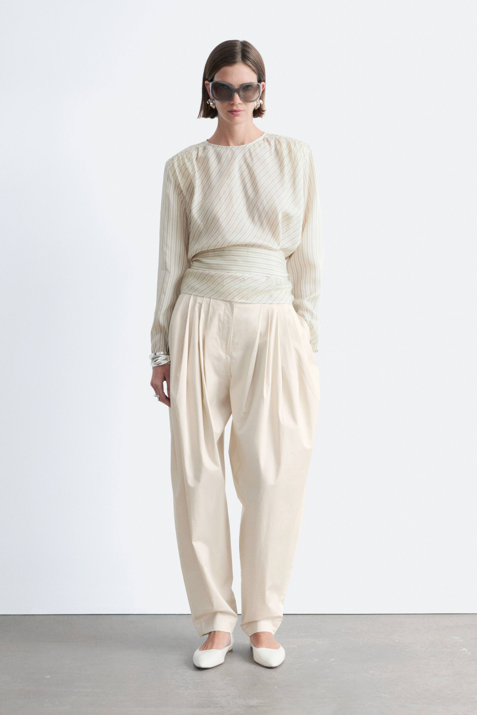 Visualizza immagine più grande: Un top beige chiaro con sottili righe diagonali presenta uno scollo a barca, maniche lunghe con spalle arricciate e una fascia in vita ampia, dello stesso tessuto, che stringe il busto. È abbinato a pantaloni beige chiaro a vita alta, caratterizzati da profonde pieghe frontali e una vestibilità rilassata e affusolata.