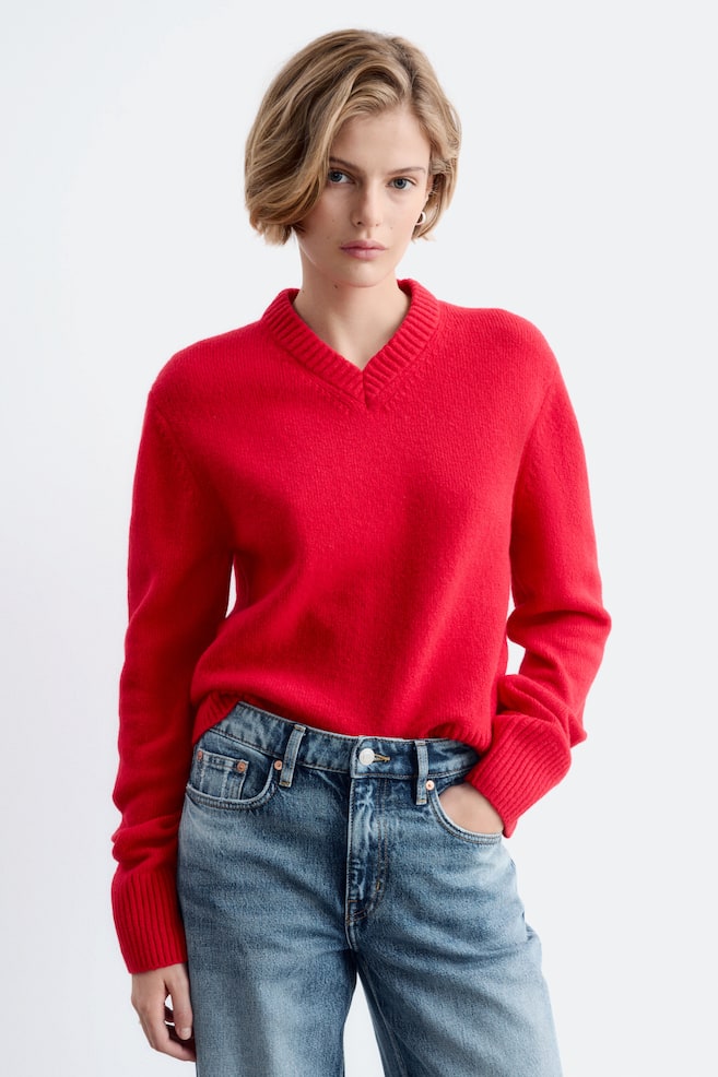 Ein roter Strickpullover mit geripptem V-Ausschnitt und gerippten BÃ¼ndchen wird von dem Model getragen. Der Pullover ist mit einer blauen Denim-Jeans kombiniert, wÃ¤hrend das Model eine Hand in die Tasche steckt. - 1