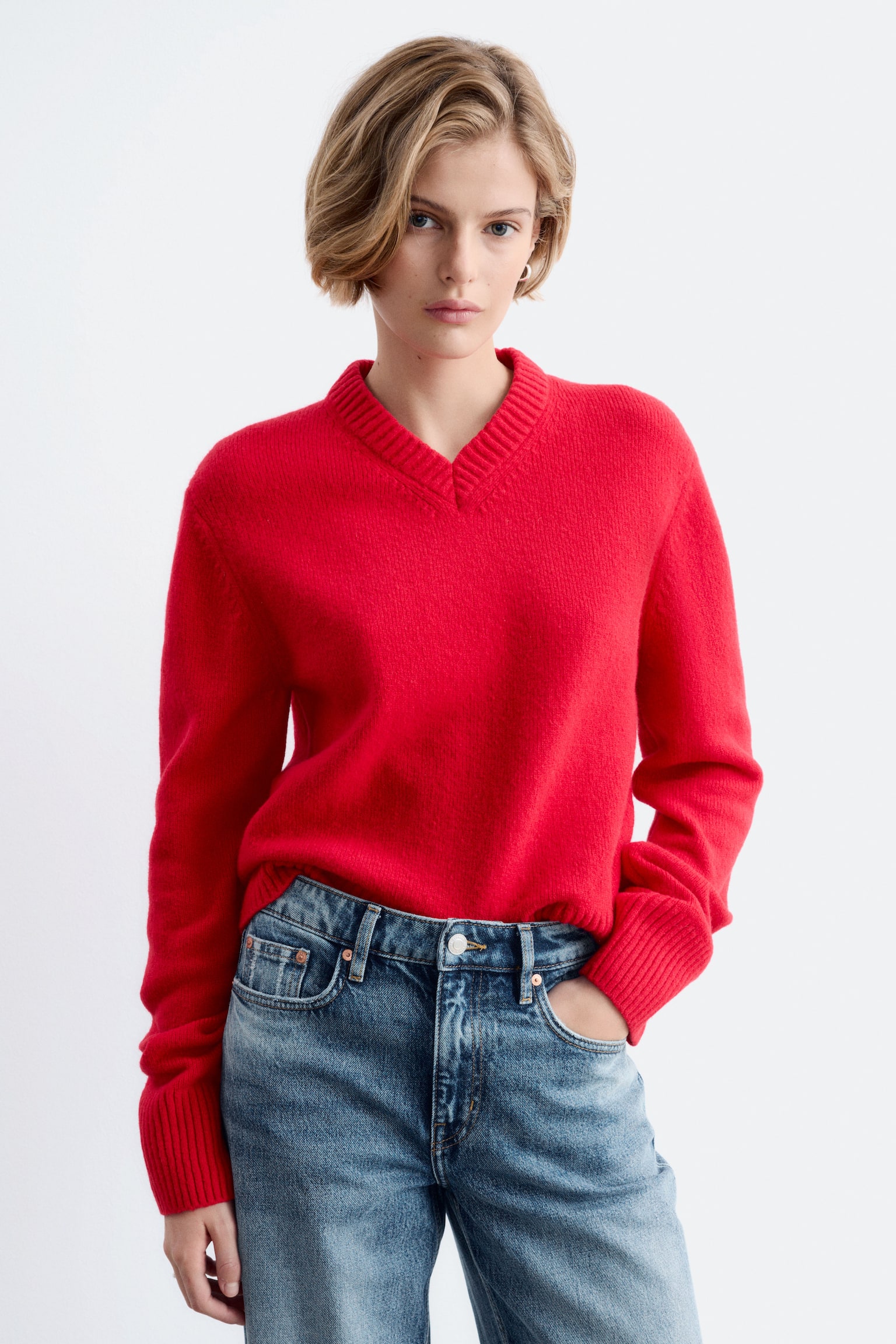 Un pull en maille rouge, doté dâun col en V côtelé et de poignets côtelés, est porté par le mannequin. Le pull est associé à un jean en denim bleu, le mannequin ayant une main dans une poche. - 1