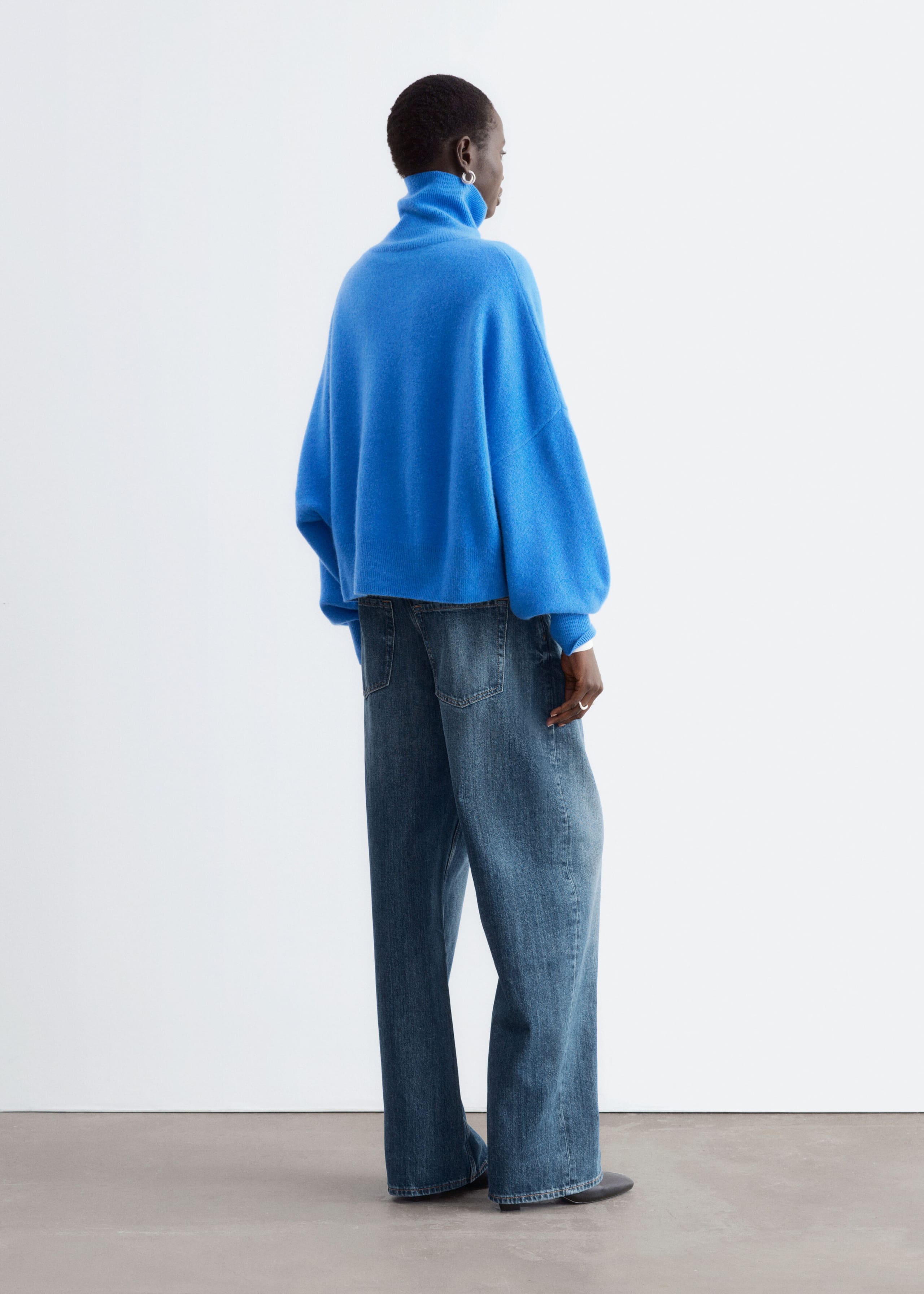 The model is wearing an oversized, bright blue wool jumper featuring a folded roll neck and dropped armholes. The jumper is styled with wide-leg blue jeans.
