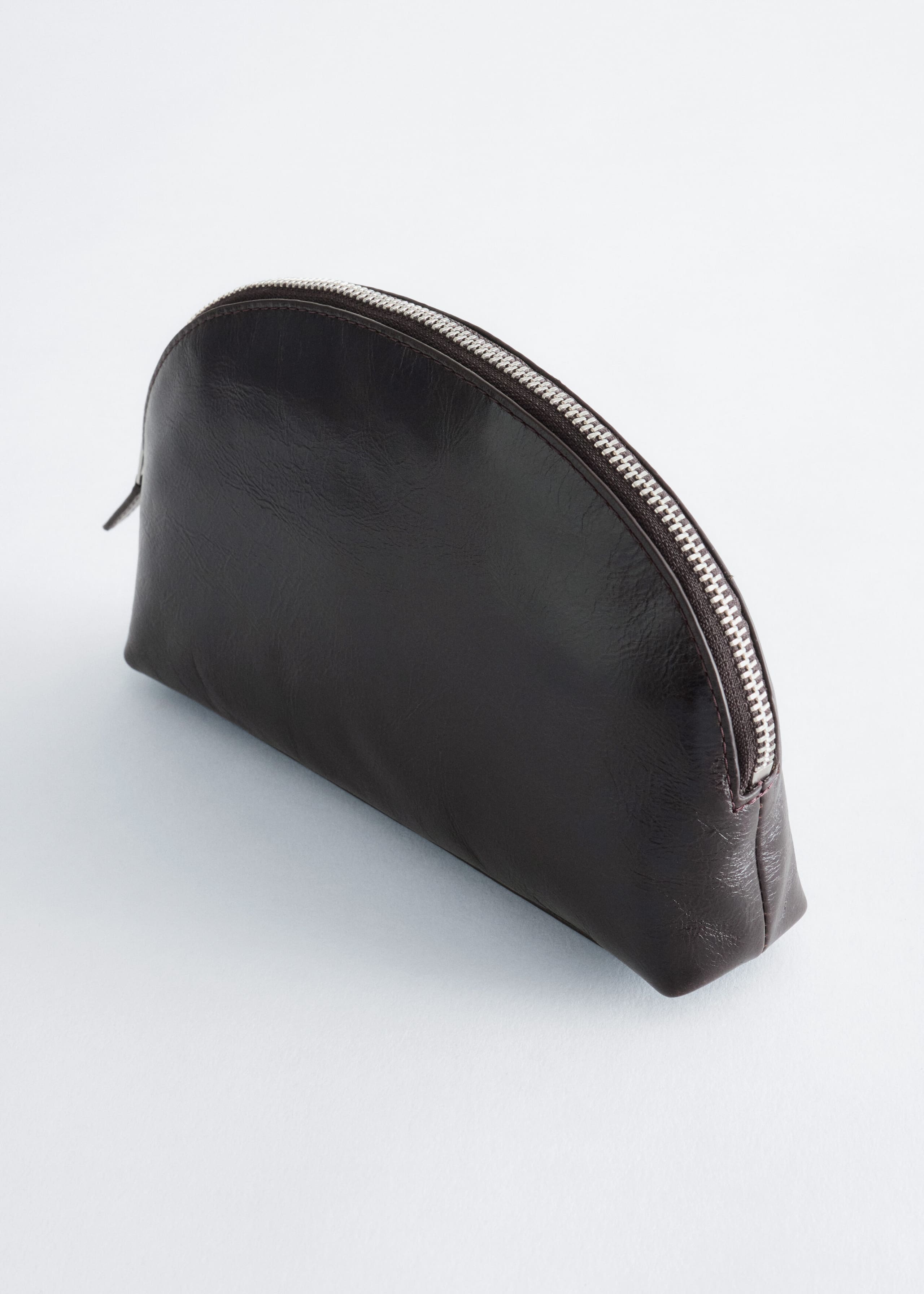 Une pochette en cuir brun foncé, dotée d'une silhouette en forme de dôme, est présentée en angle trois-quarts, mettant en valeur sa surface texturée semi-brillante. Une fermeture éclair métallique ton argent court le long du bord supérieur incurvé de la pochette.