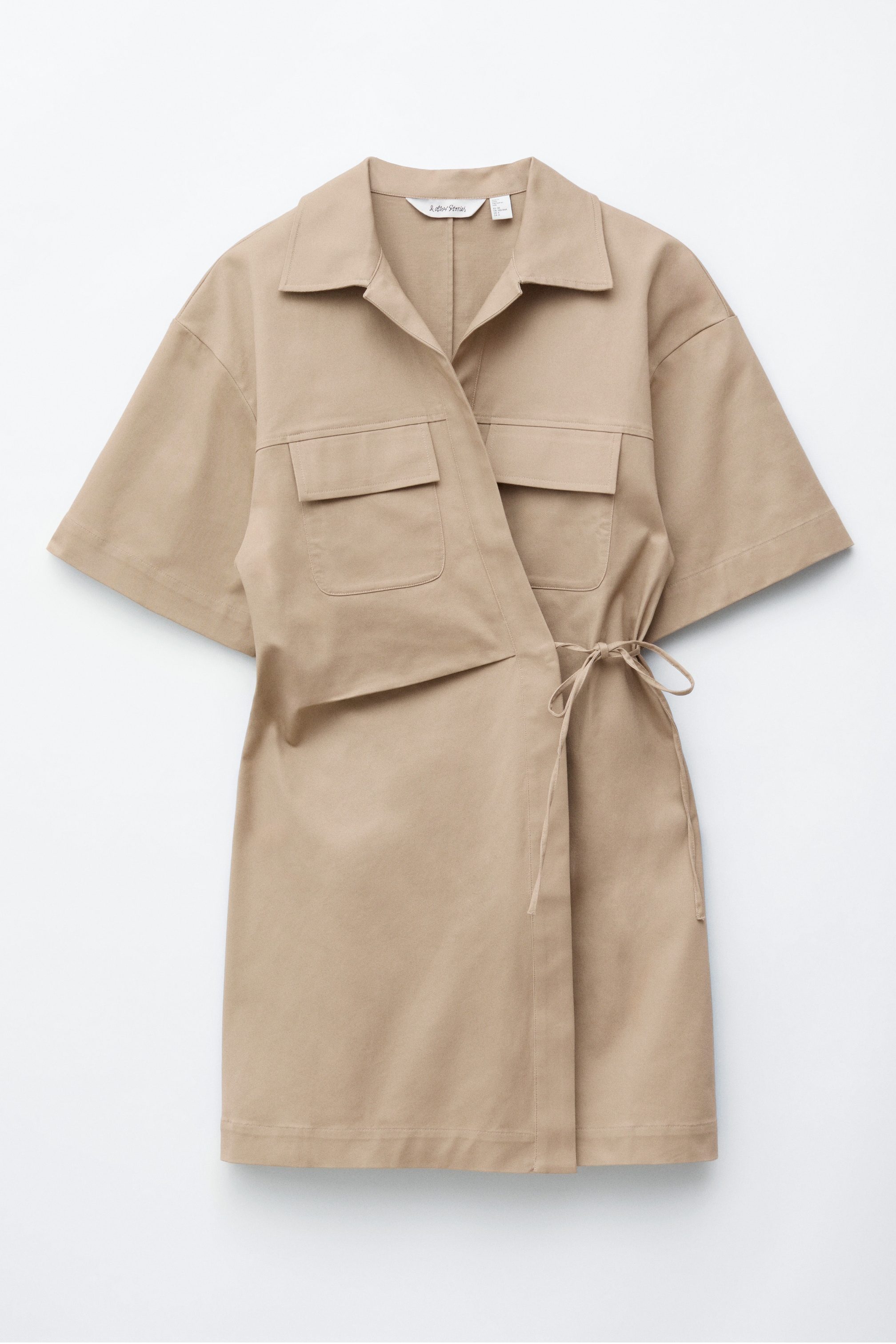 Visualizza immagine più grande: Un abito a portafoglio beige è visto di fronte, caratterizzato da un colletto a punta e maniche corte e ampie. Due grandi tasche con patta sono visibili sul petto, e un sottile nastro dello stesso tessuto si annoda sul fianco.