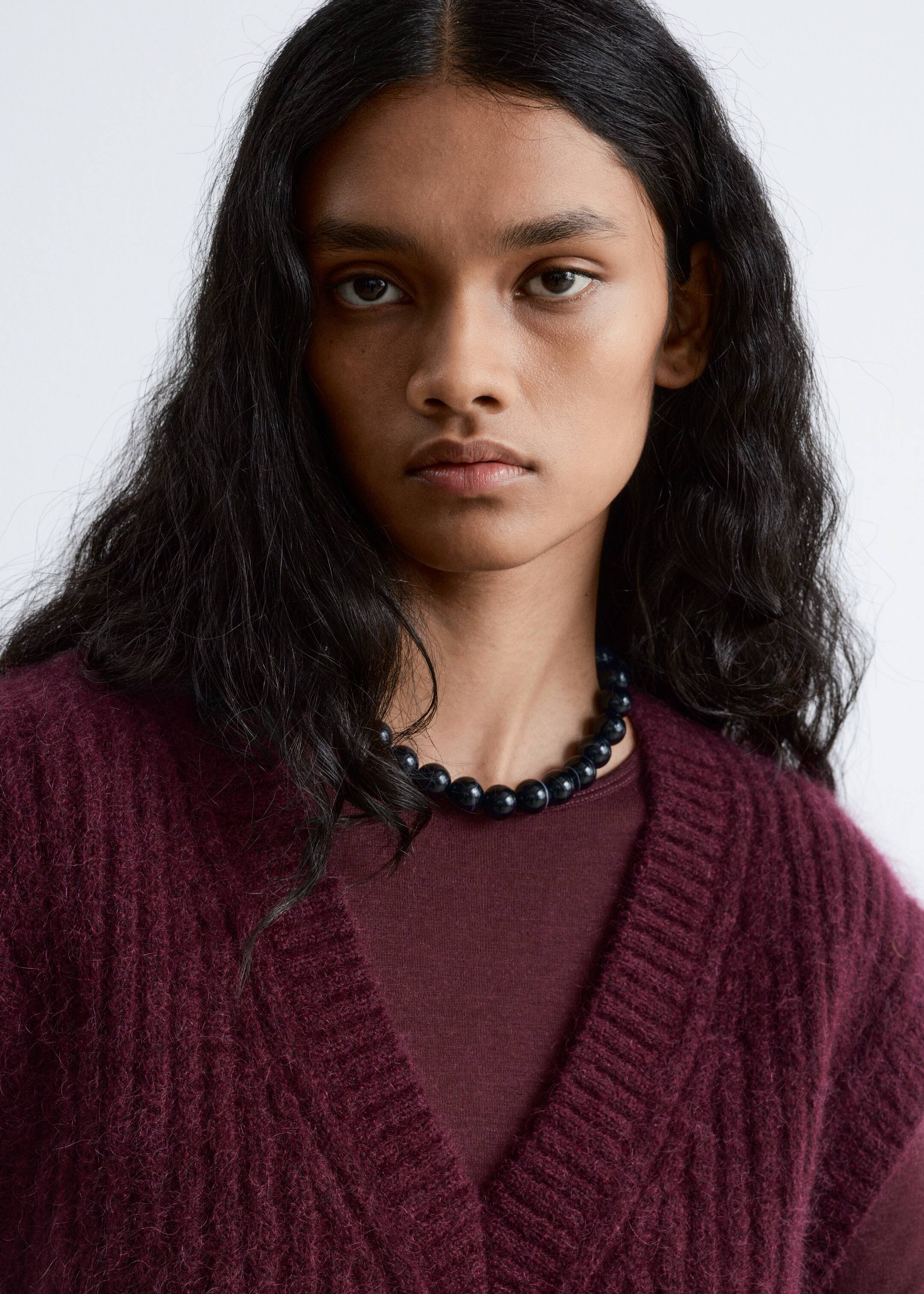Größeres Bild ansehen: Eine burgunderrote V-Ausschnitt-Strickjacke mit flauschiger Textur und geripptem Besatz entlang des nach vorne gerichteten V-Ausschnitts wird über einem passenden burgunderroten Rundhalsoberteil getragen. Eine dunkle Perlenkette mit großen runden Perlen wird um den Hals getragen und ruht über dem Ausschnitt des Oberteils.