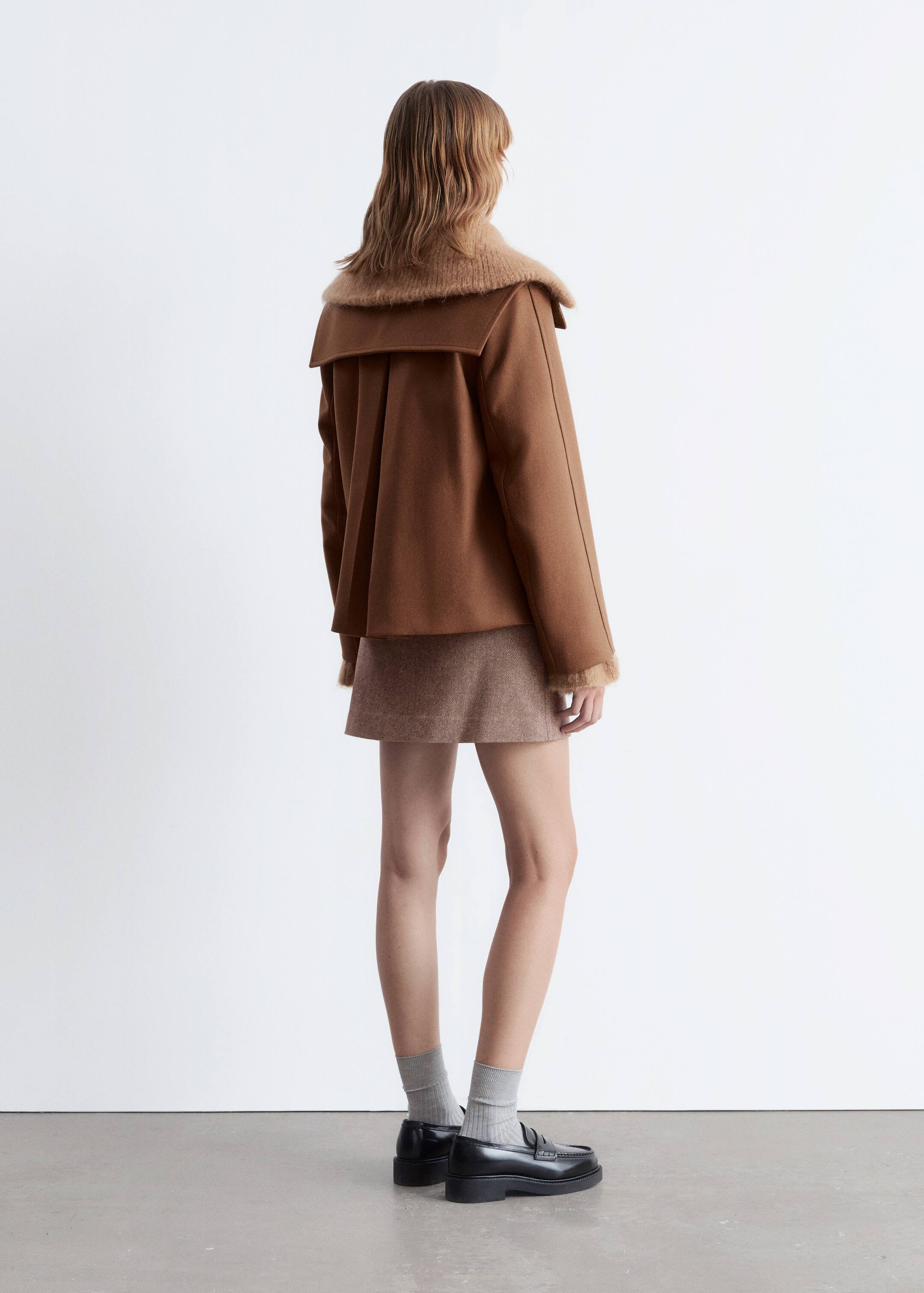 Agrandir l'image: Un manteau caban marron en ligne A est porté par le mannequin, qui se tient dos au spectateur. Le manteau présente un col marin carré proéminent et un pli creux à l'ourlet arrière. Un pull à col montant marron clair avec une finition texturée est superposé sous le manteau, avec son col et ses poignets visiblement étendus.