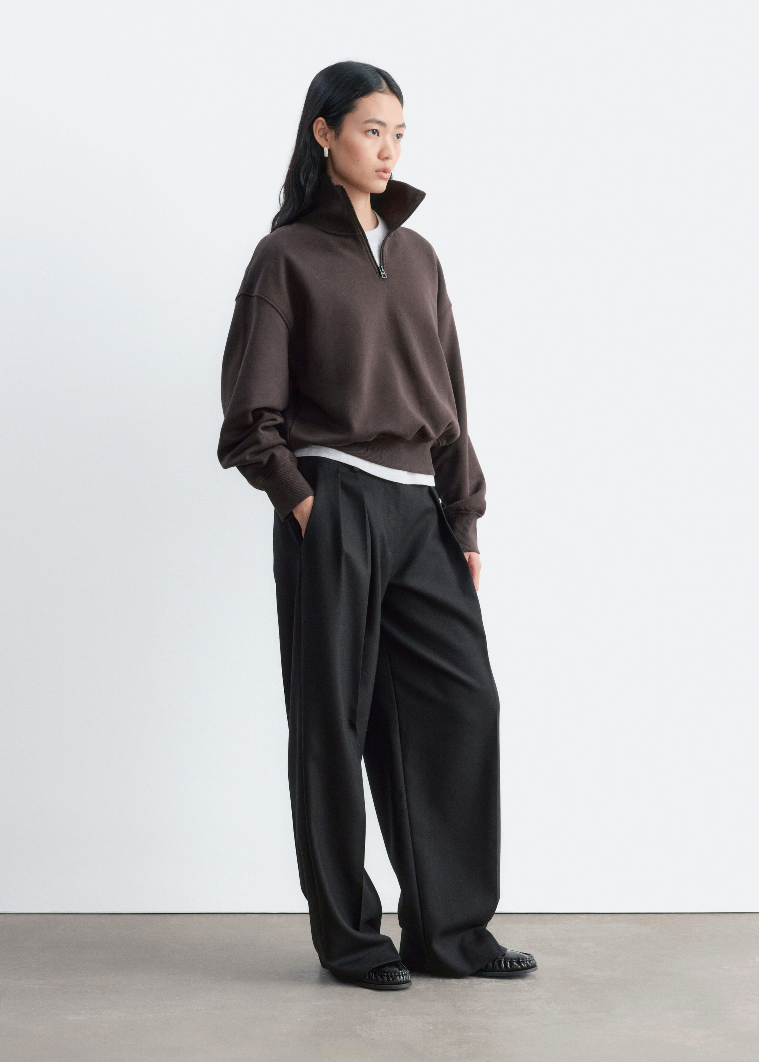 A dark brown cotton jersey sweatshirt with a half-zip collar and ribbed trims is being worn by the model. The sweatshirt is layered over a white T-shirt and styled with wide-leg black trousers.